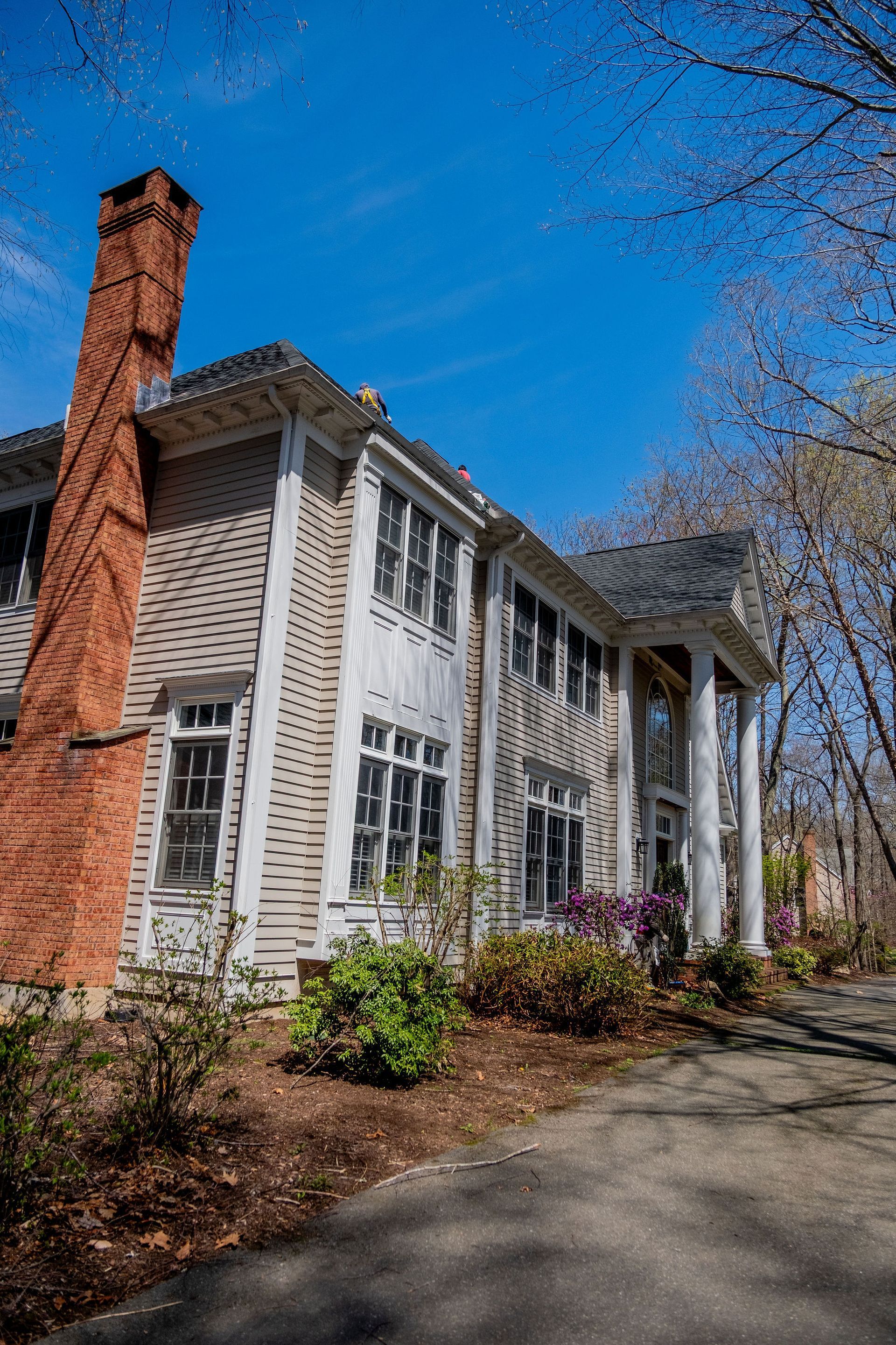 Two-story light gray house with tall brick chimney, white columns, and blue sky.
