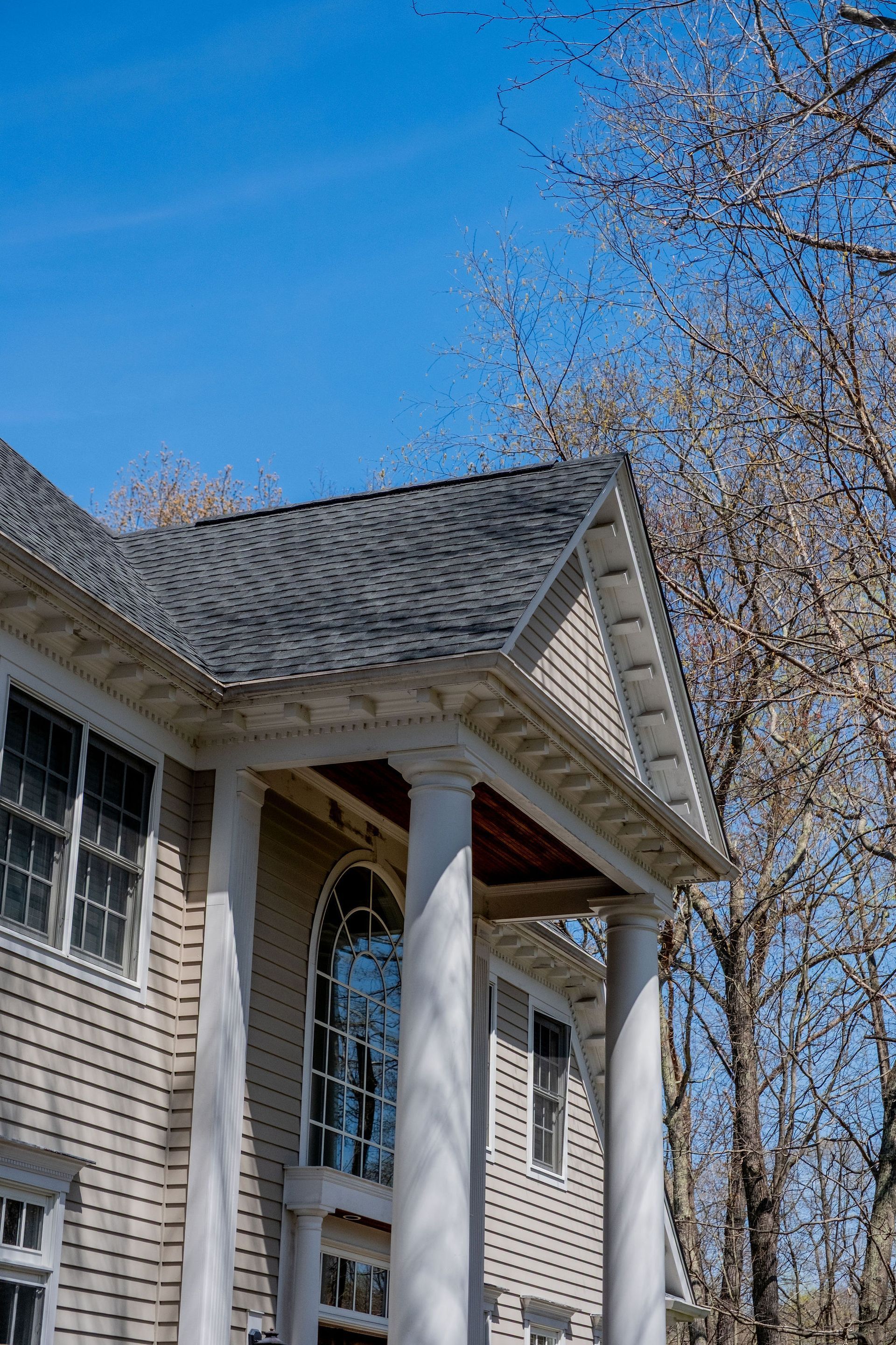 Beige house with white columns and dark gray roof against a blue sky, tall tree in the background.