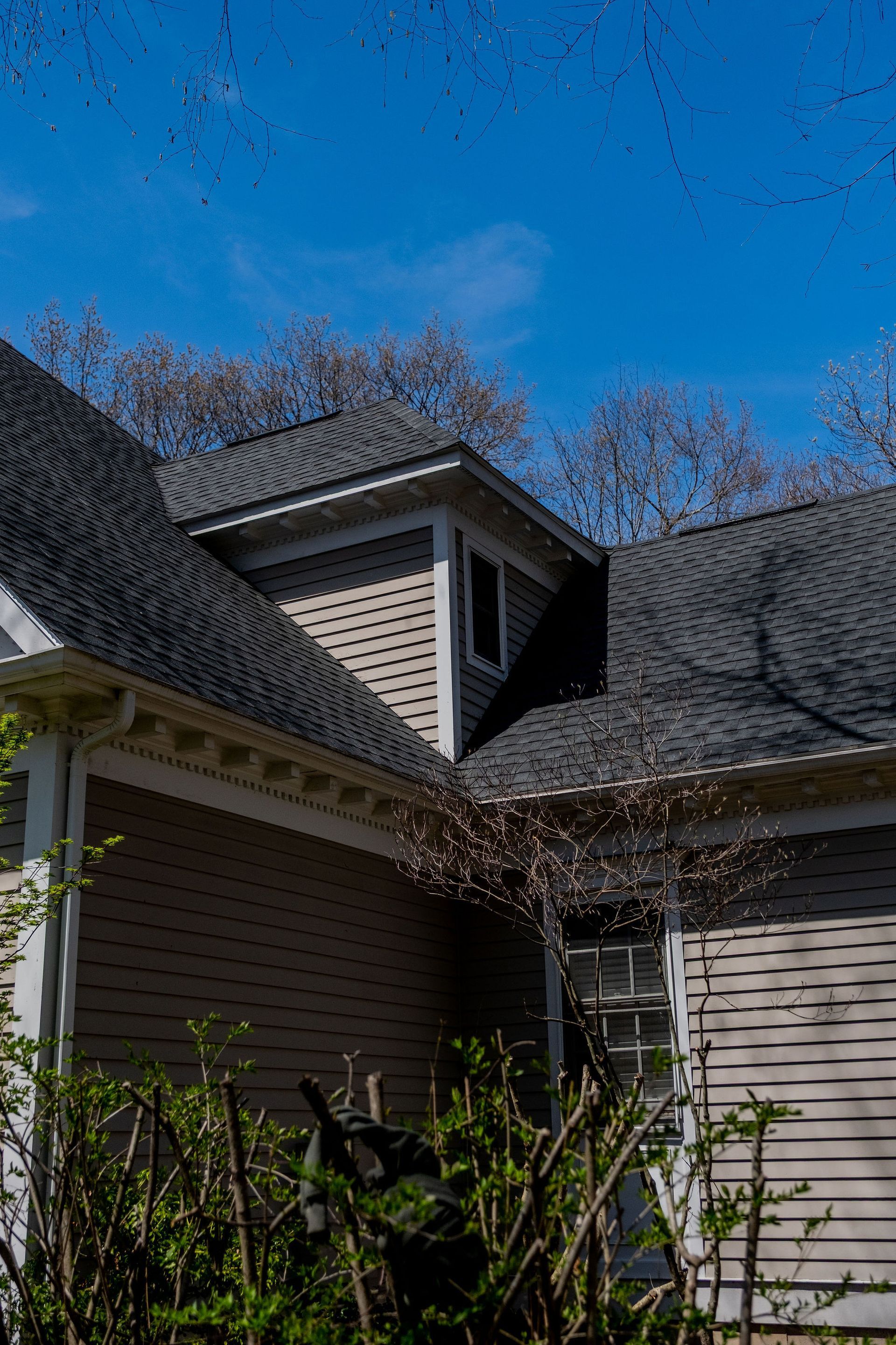 Gray house exterior with dark roof, dormer, and trees against a blue sky.
