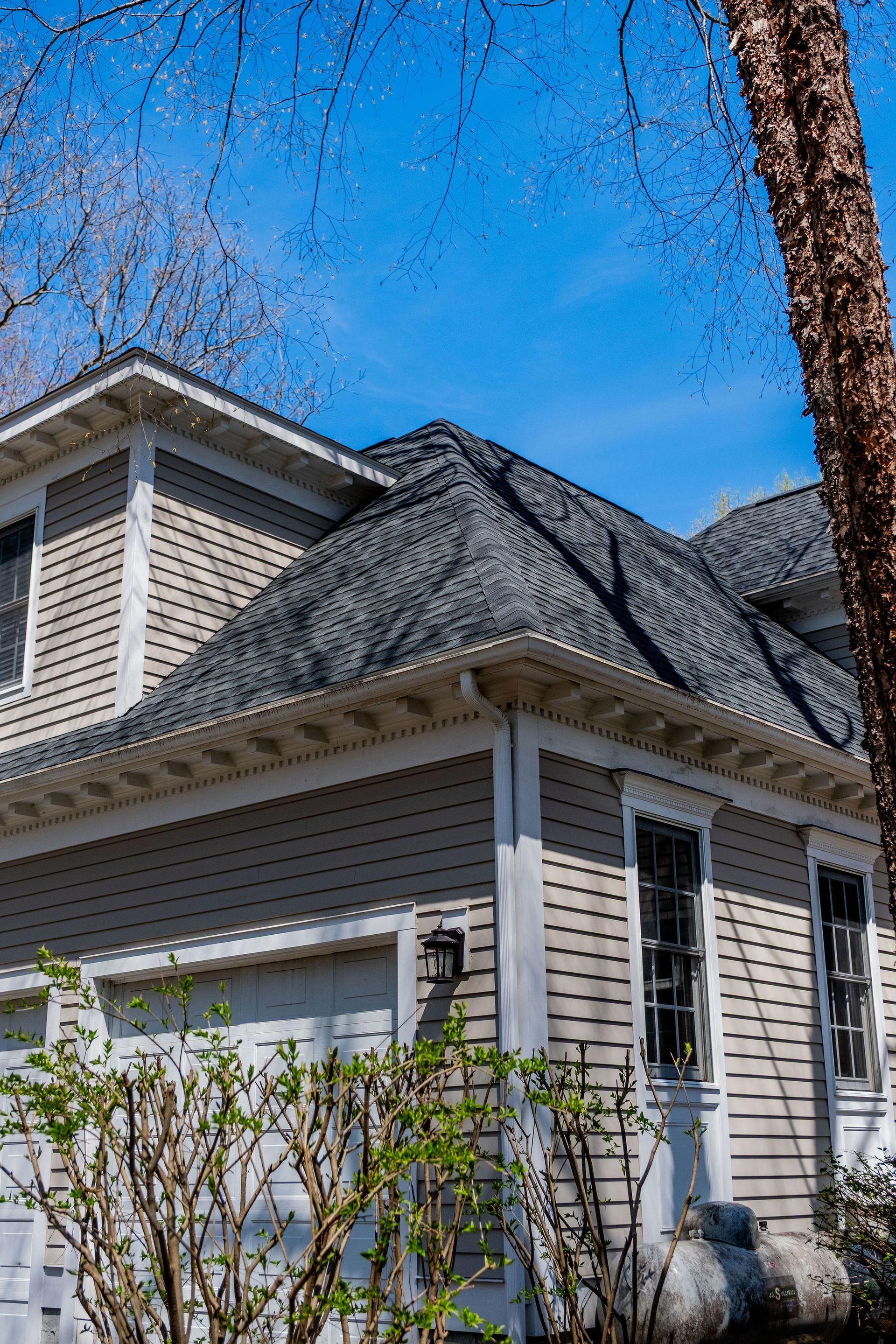 Two-story beige house with a dark gray roof. The house has a garage and a tree to the right, blue sky in the background.