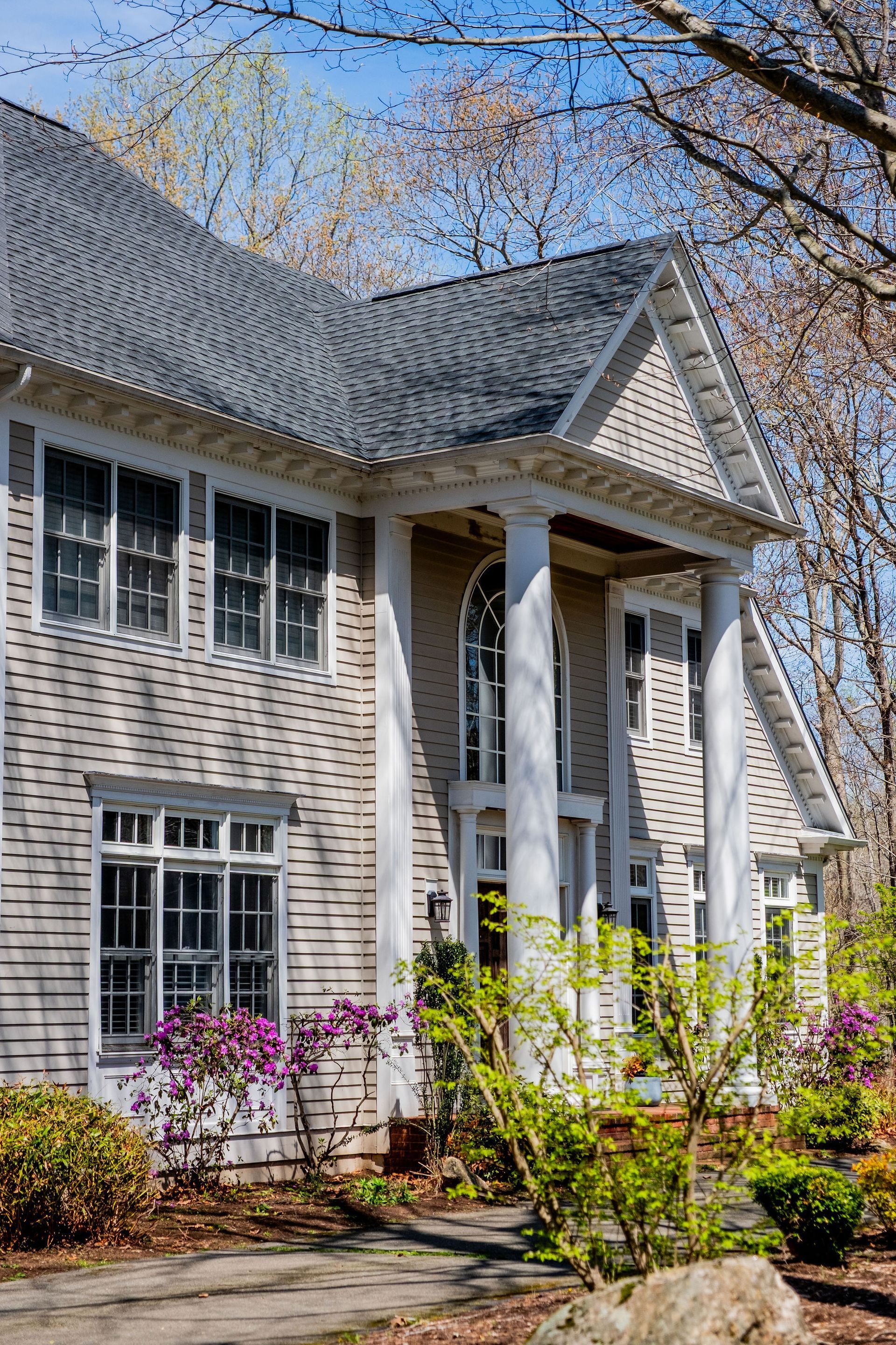 Two-story house with gray shingles, white columns, and windows. Flowering bushes and trees in the yard.