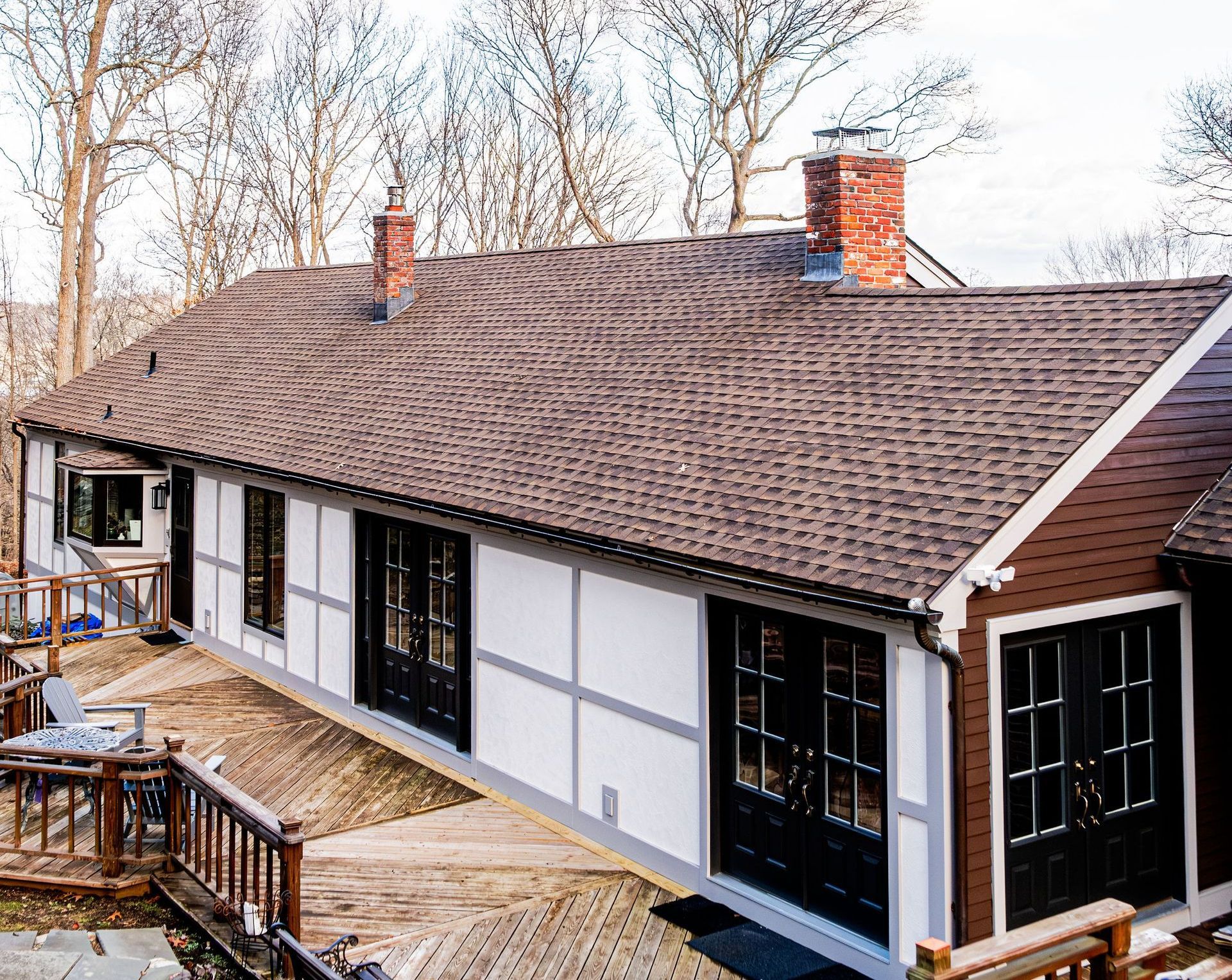 A white and brown Tudor-style house with black doors and a wooden deck. The roof has two chimneys.