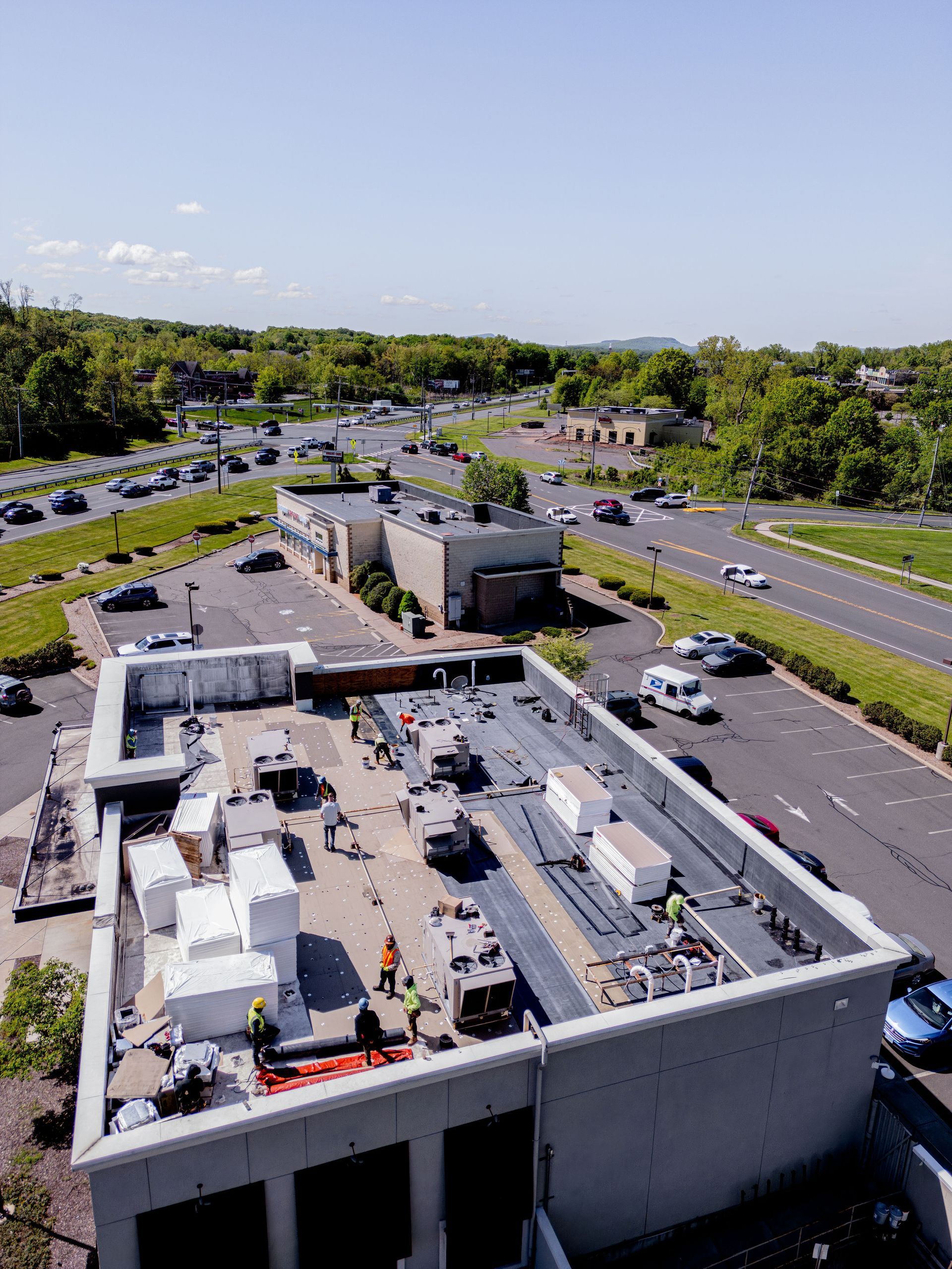 Construction workers on a flat roof next to a road, trees, and buildings; sunny day.