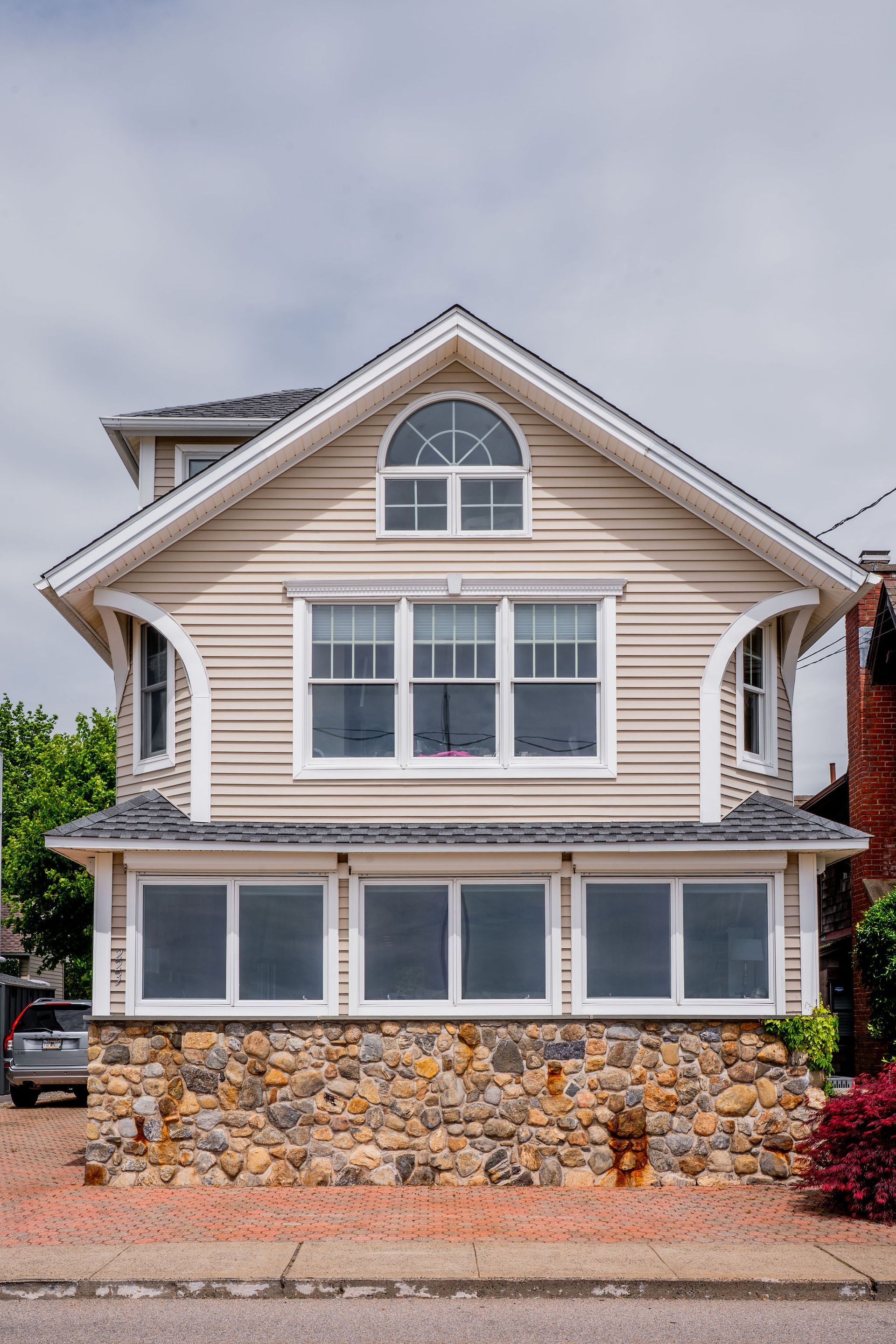 Two-story beige house with stone foundation, multiple windows, and a curved roof design under a cloudy sky.