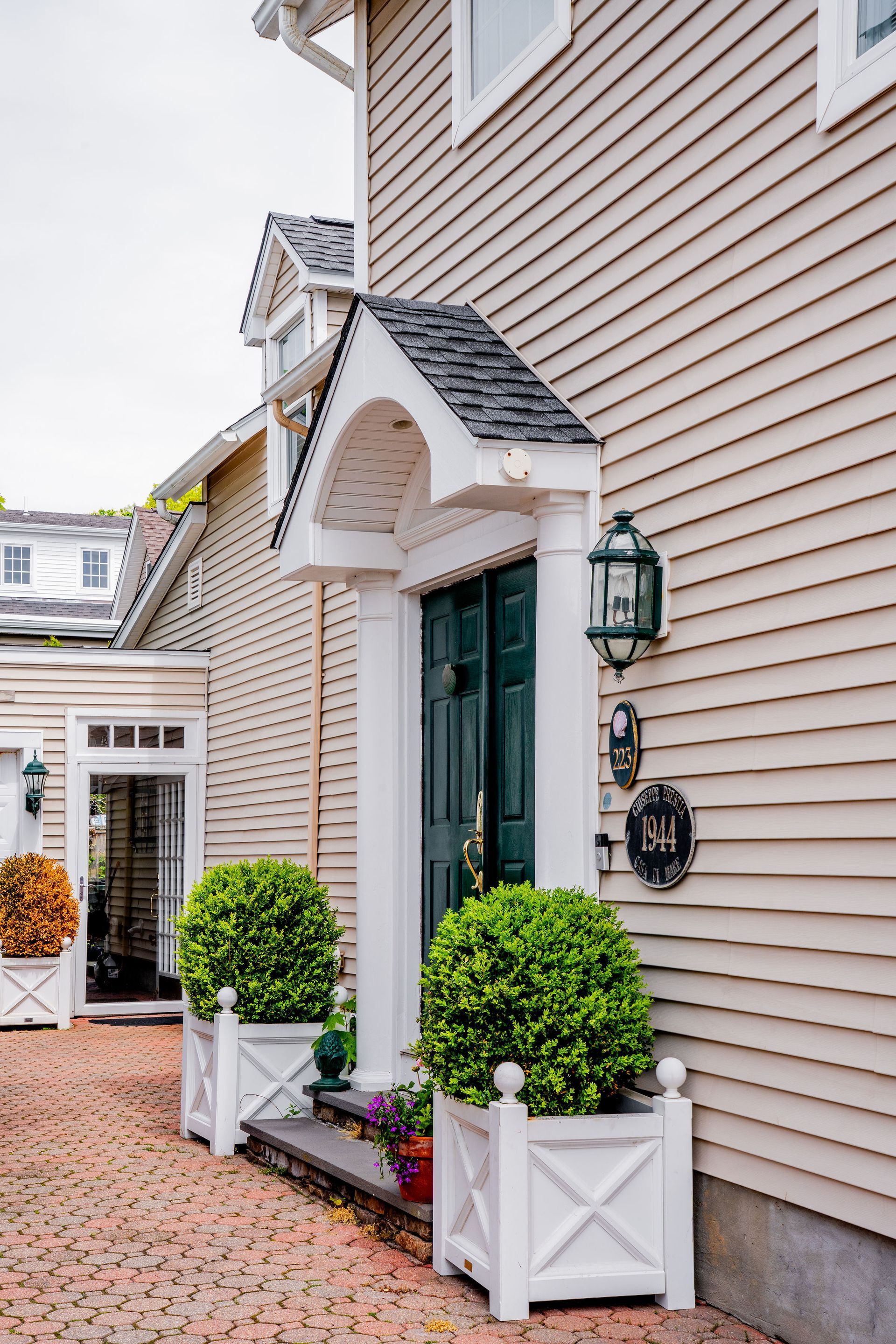 Beige house exterior with white trim, green door, brick walkway, and potted shrubs on either side