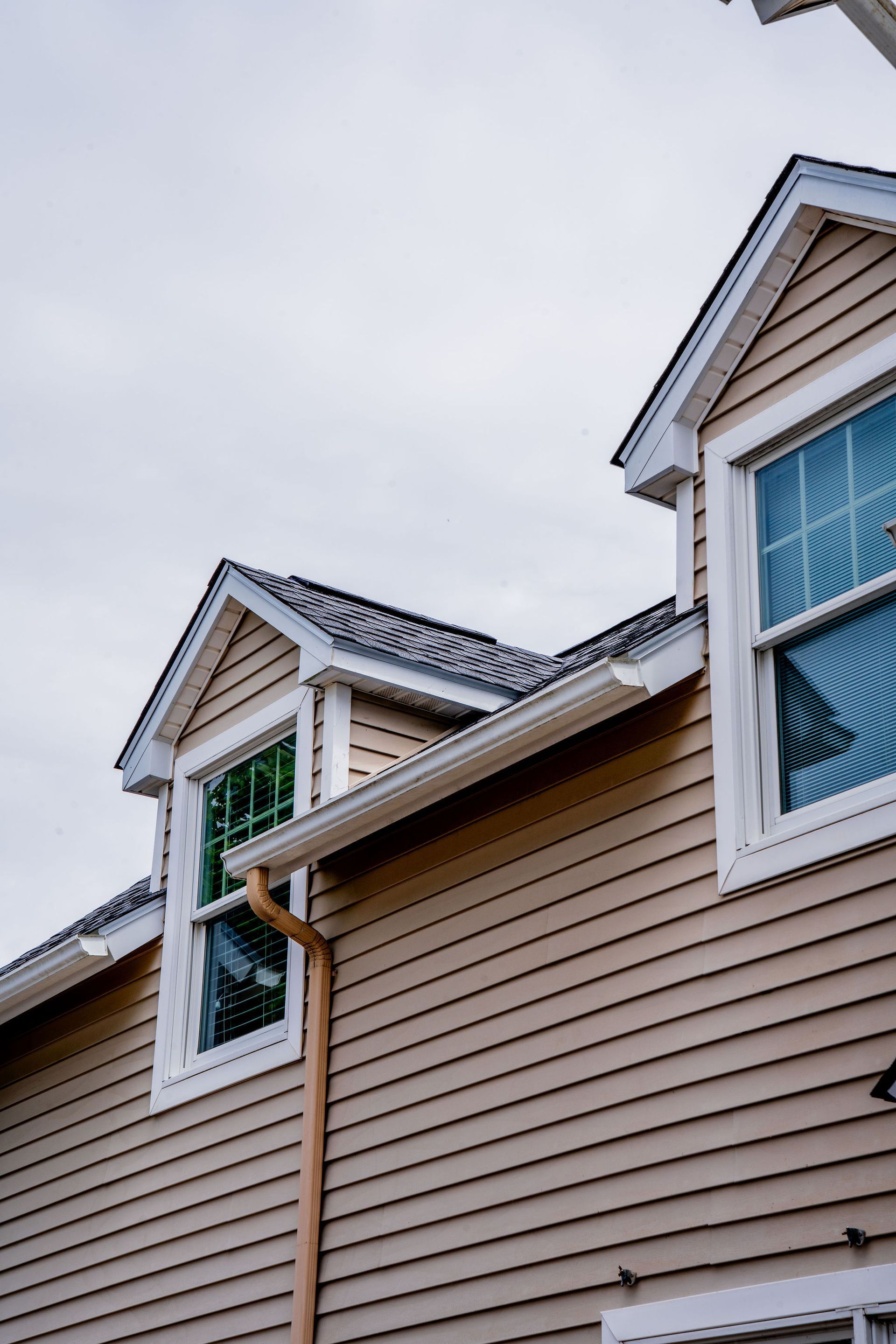 Tan siding with white-framed windows, two dormers, and tan gutters against a cloudy sky.