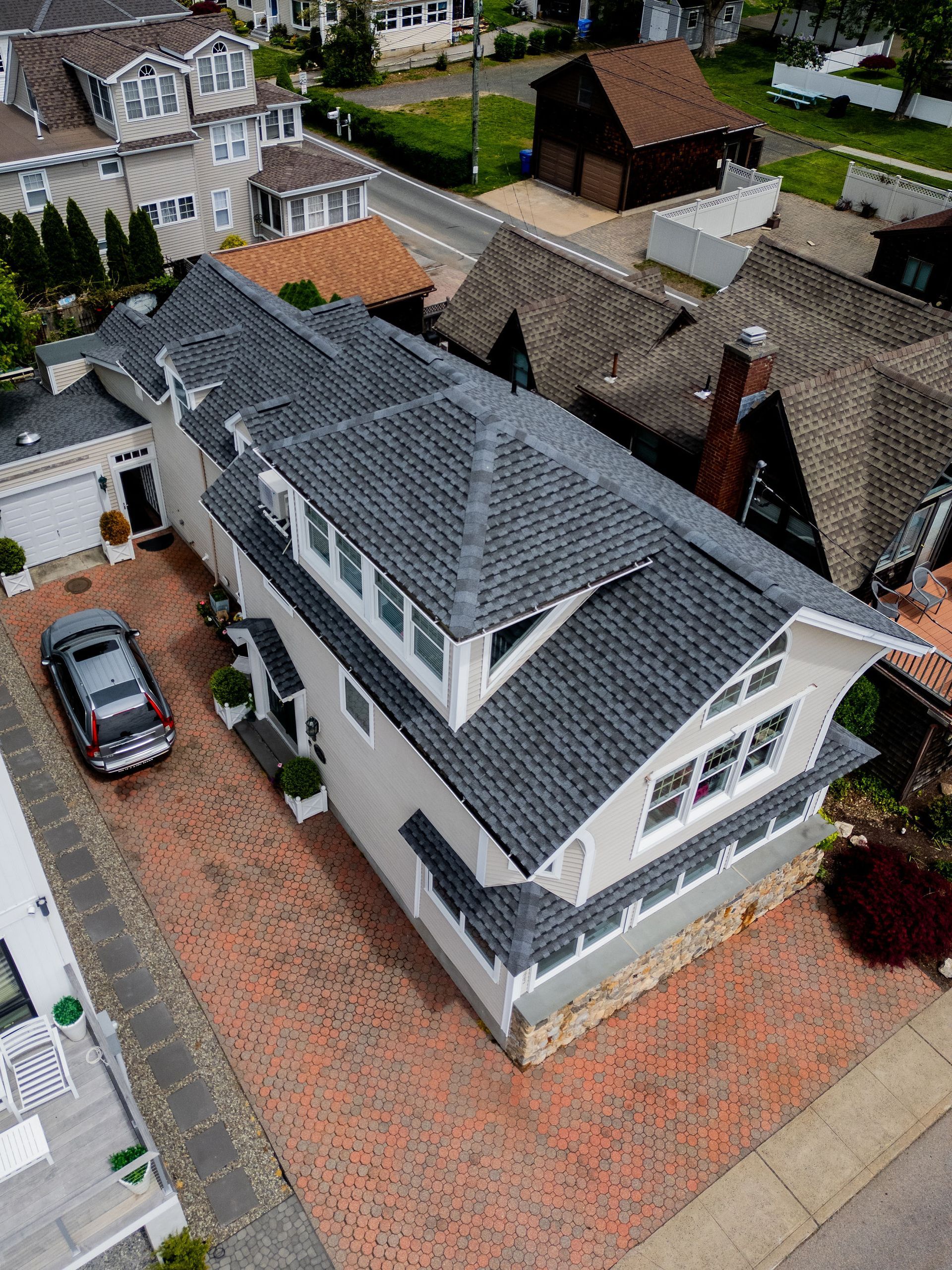 Aerial view of a two-story house with a brick driveway and dark gray roof. A car is parked on the driveway.
