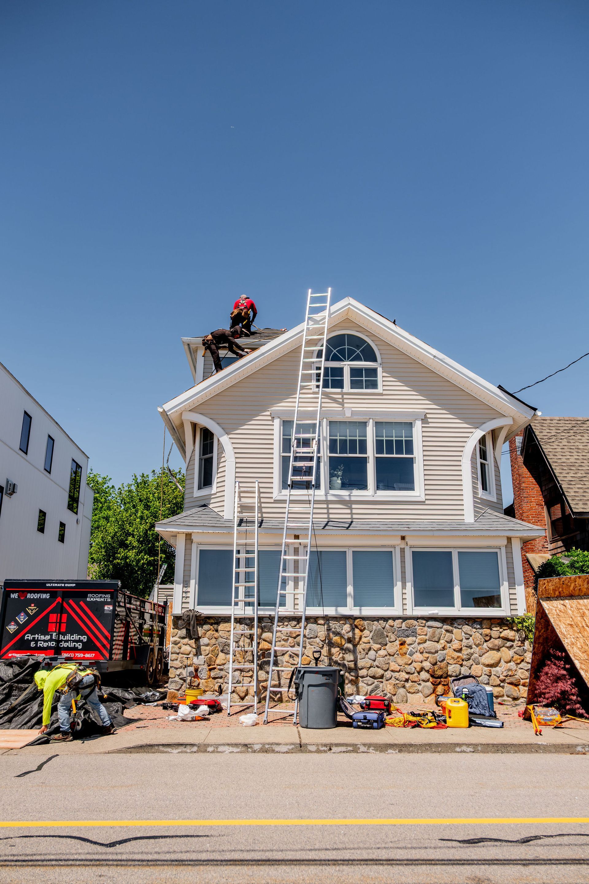 Two-story house with ladder and workers. Roof repair in progress; sunny day with clear blue sky.