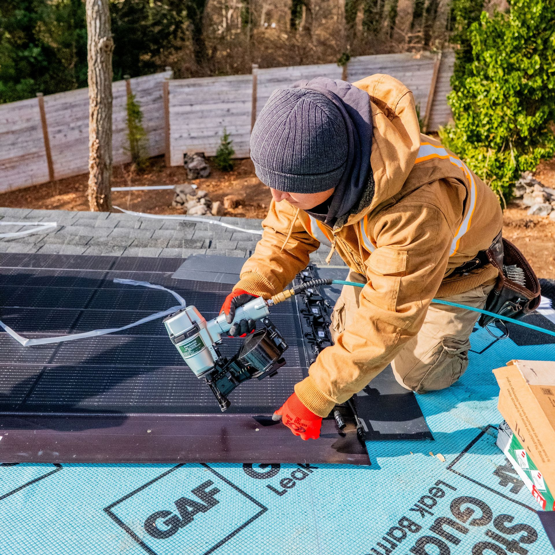 Roofer kneels, using a nail gun on a roof. Brown jacket, red gloves, gray hat. GAF underlayment visible.