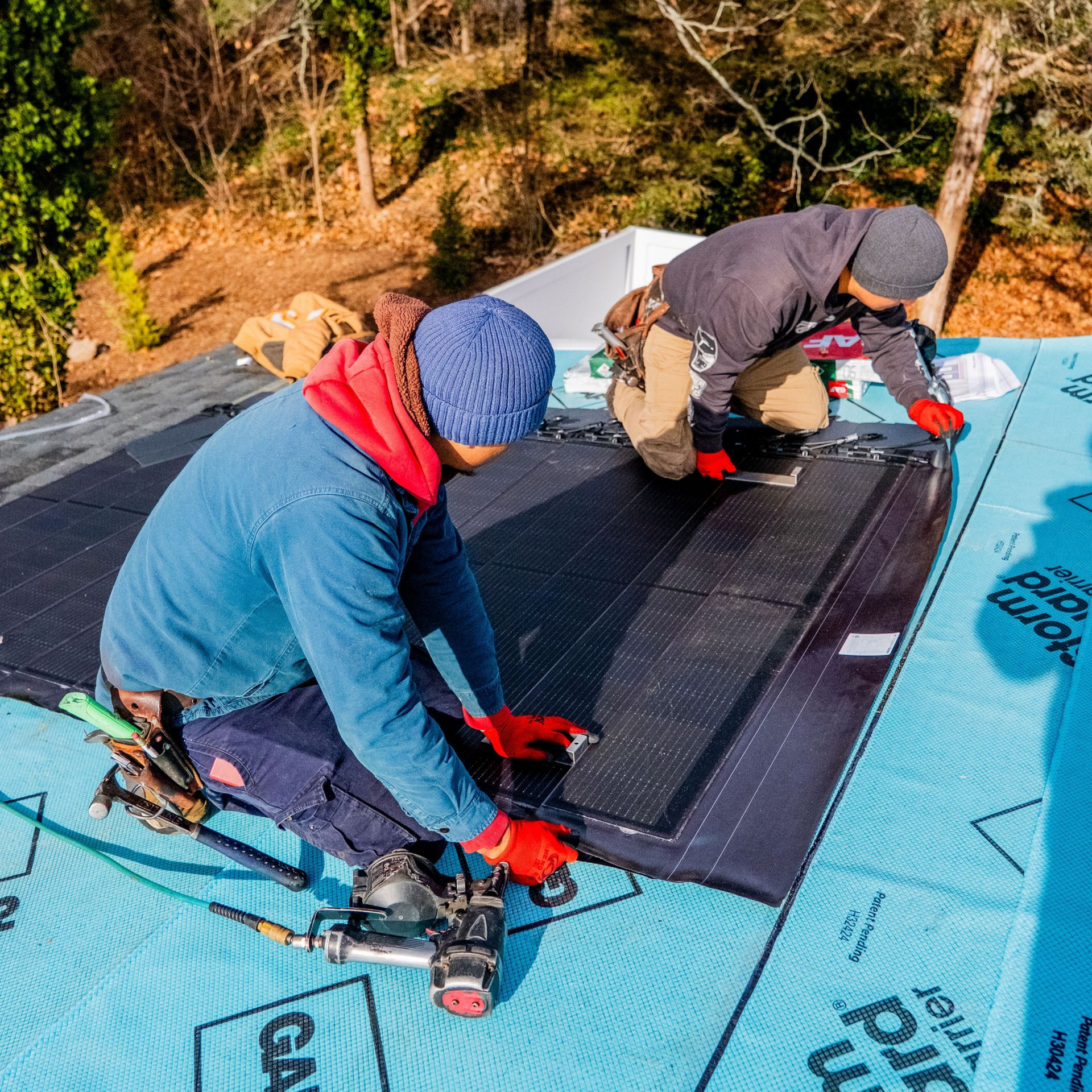 Two workers installing solar panels on a rooftop. They are wearing gloves and beanies.