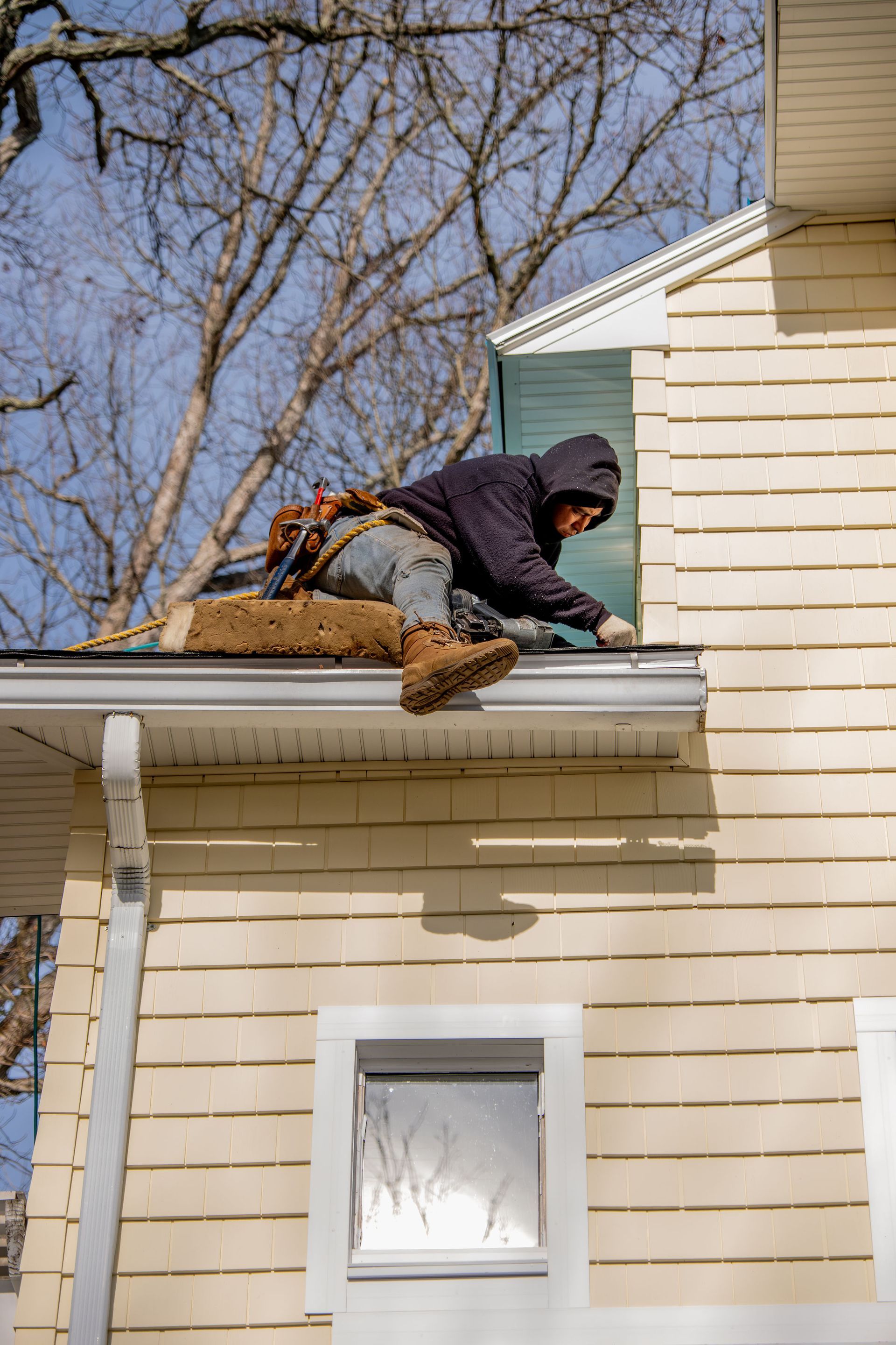 Person in hooded jacket works on gutter of a house; sunny day, bare tree in background.