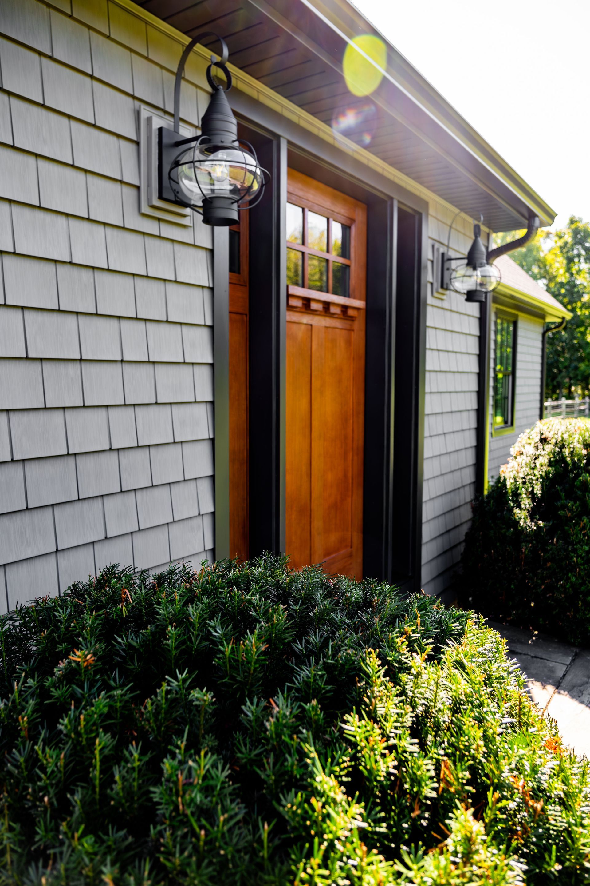 Wooden door with black trim, gray shake siding, and exterior light fixtures. Green shrub in foreground.