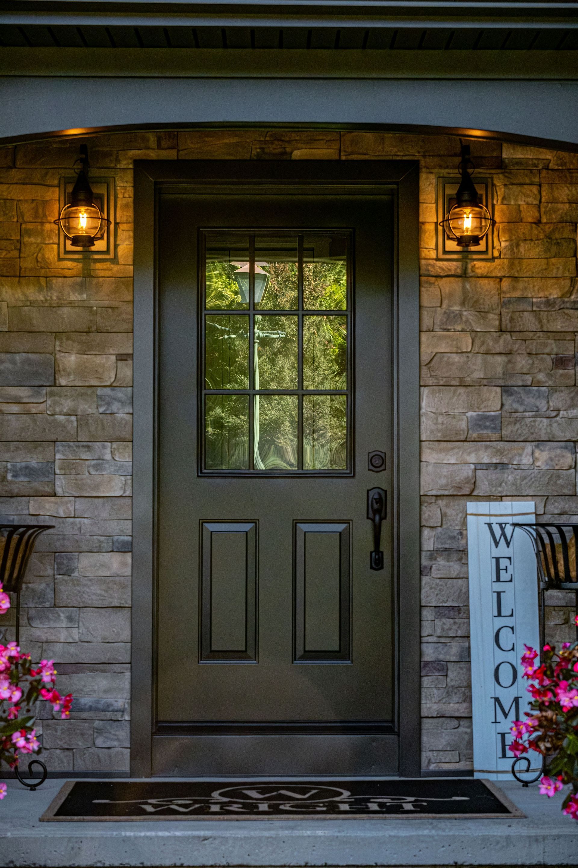 Dark brown front door with glass window, flanked by lamps, set in a stone wall. Welcome sign and flowers visible.