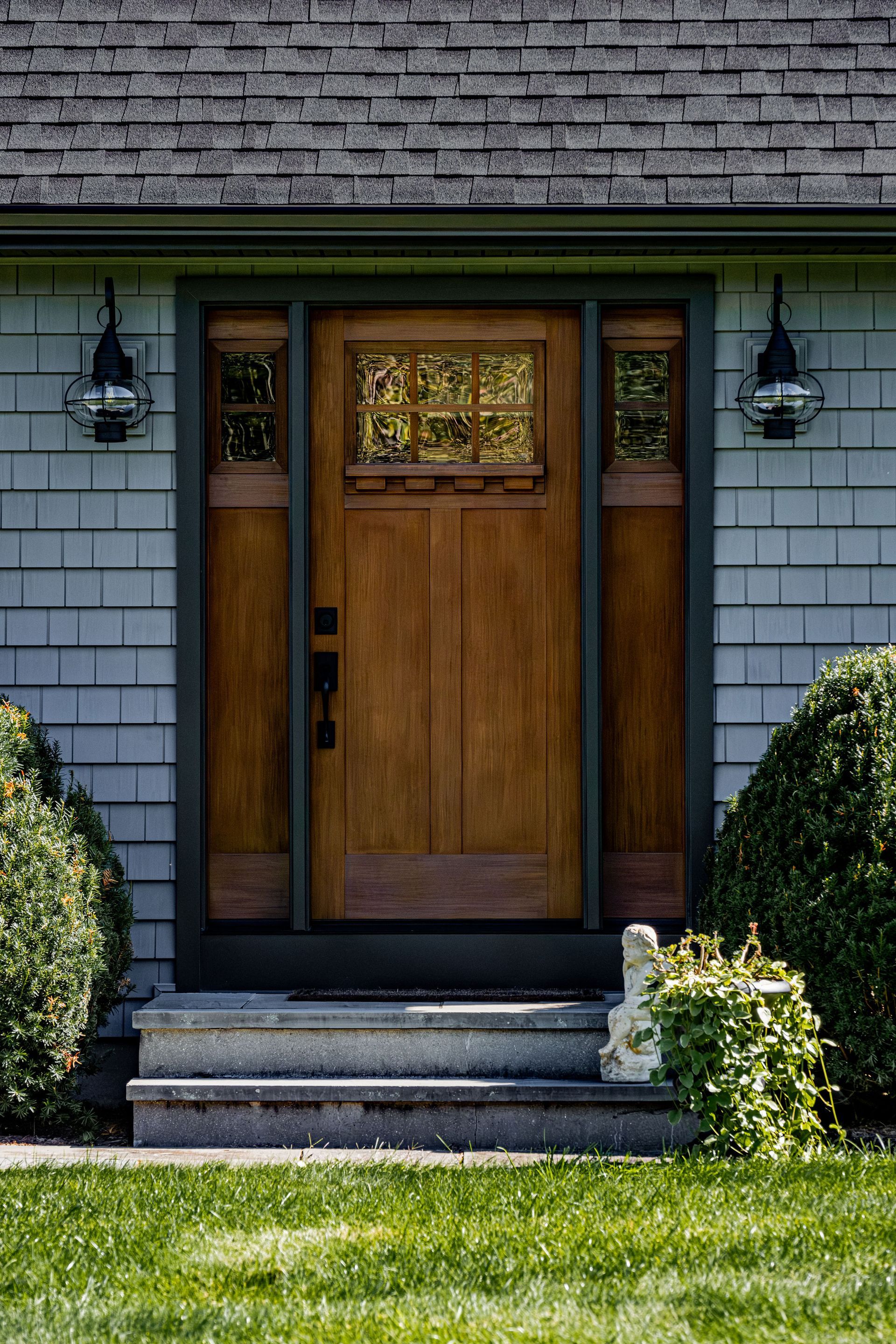 Wooden front door with sidelights, steps, and lanterns on a gray shingled house.
