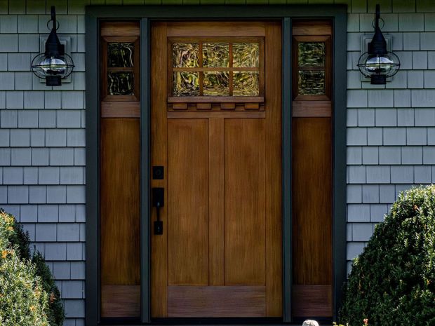 Wooden front door with sidelights and transom window, flanked by outdoor lanterns, on a shingled house.
