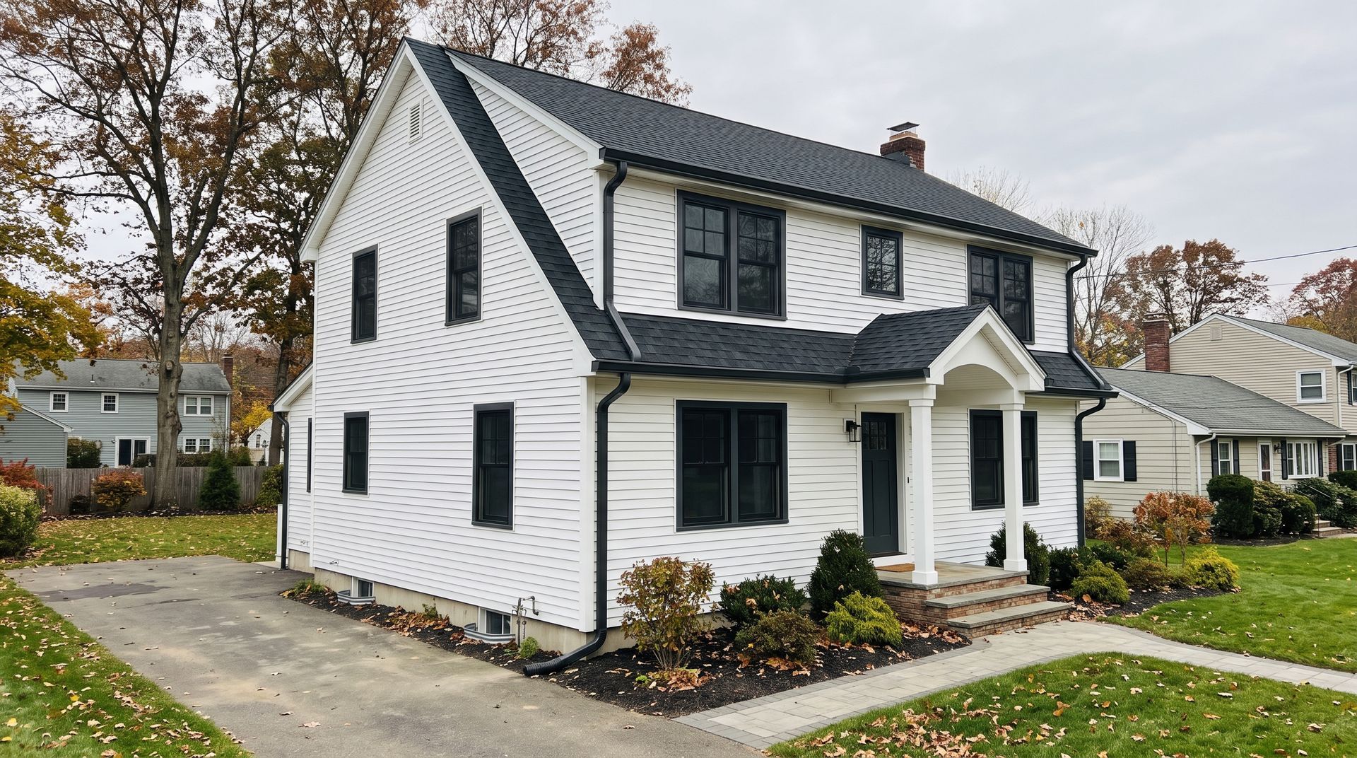 White two-story suburban house with black shutters, front porch, and autumn trees along a driveway