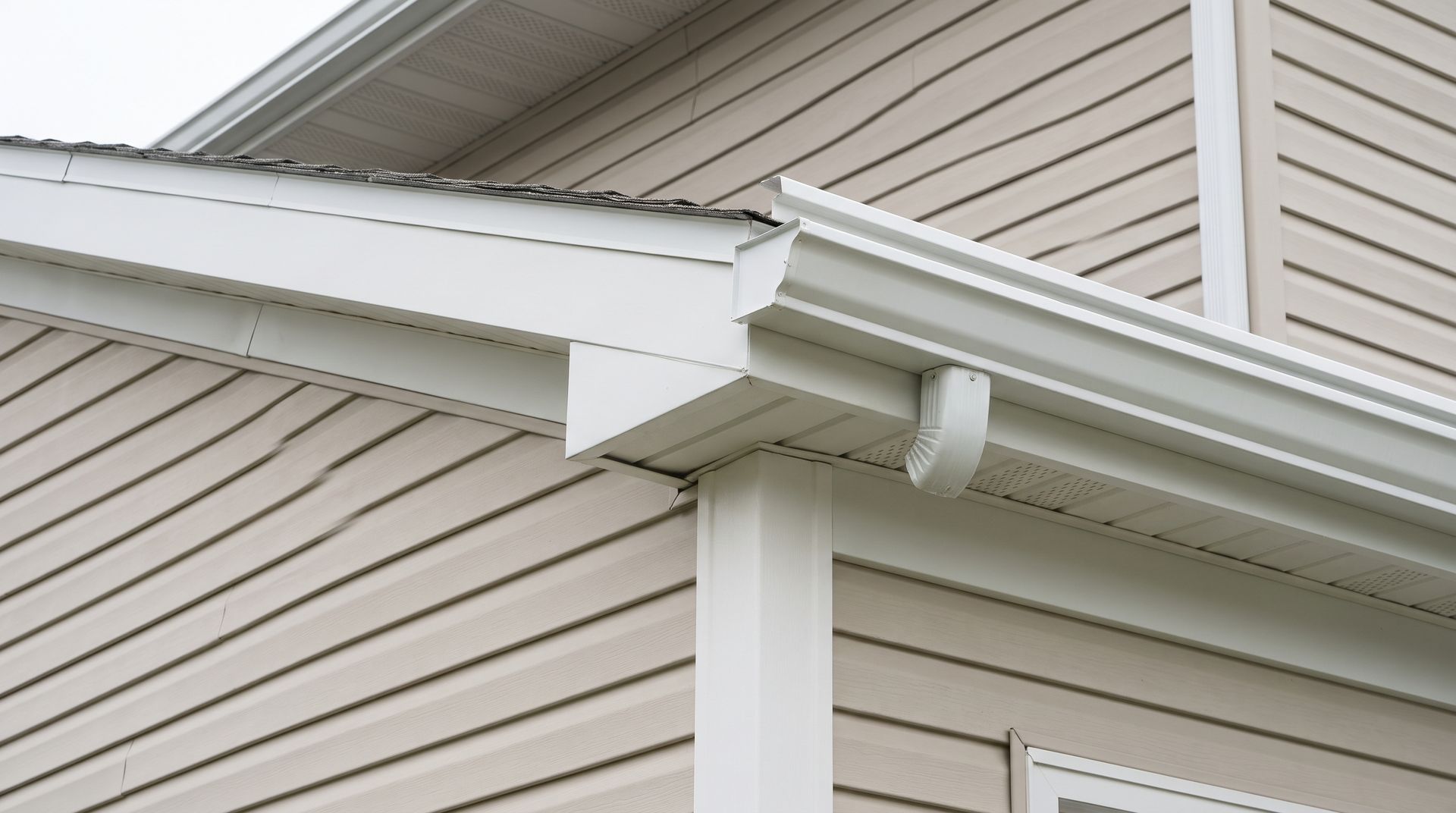 White house gutter and downspout under a beige vinyl-sided roofline