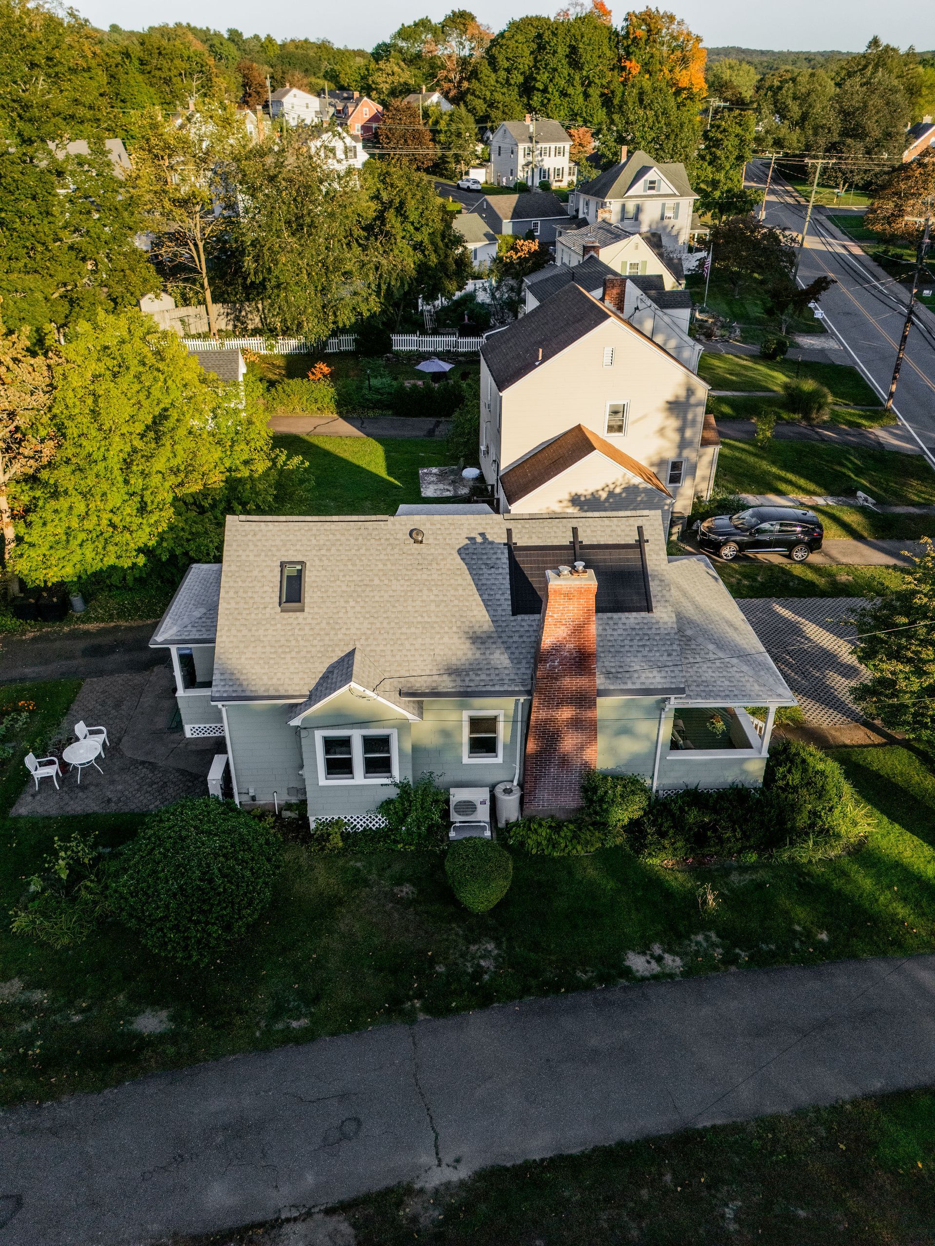 An aerial view of a light green house with a brick chimney, surrounded by trees and other houses.