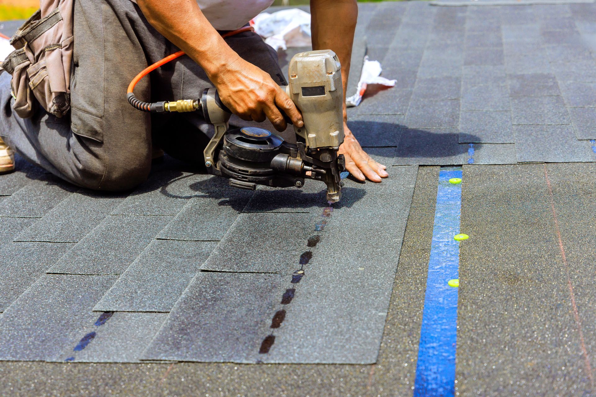 A construction worker uses a pneumatic nail gun to install asphalt shingles on a residential roof.