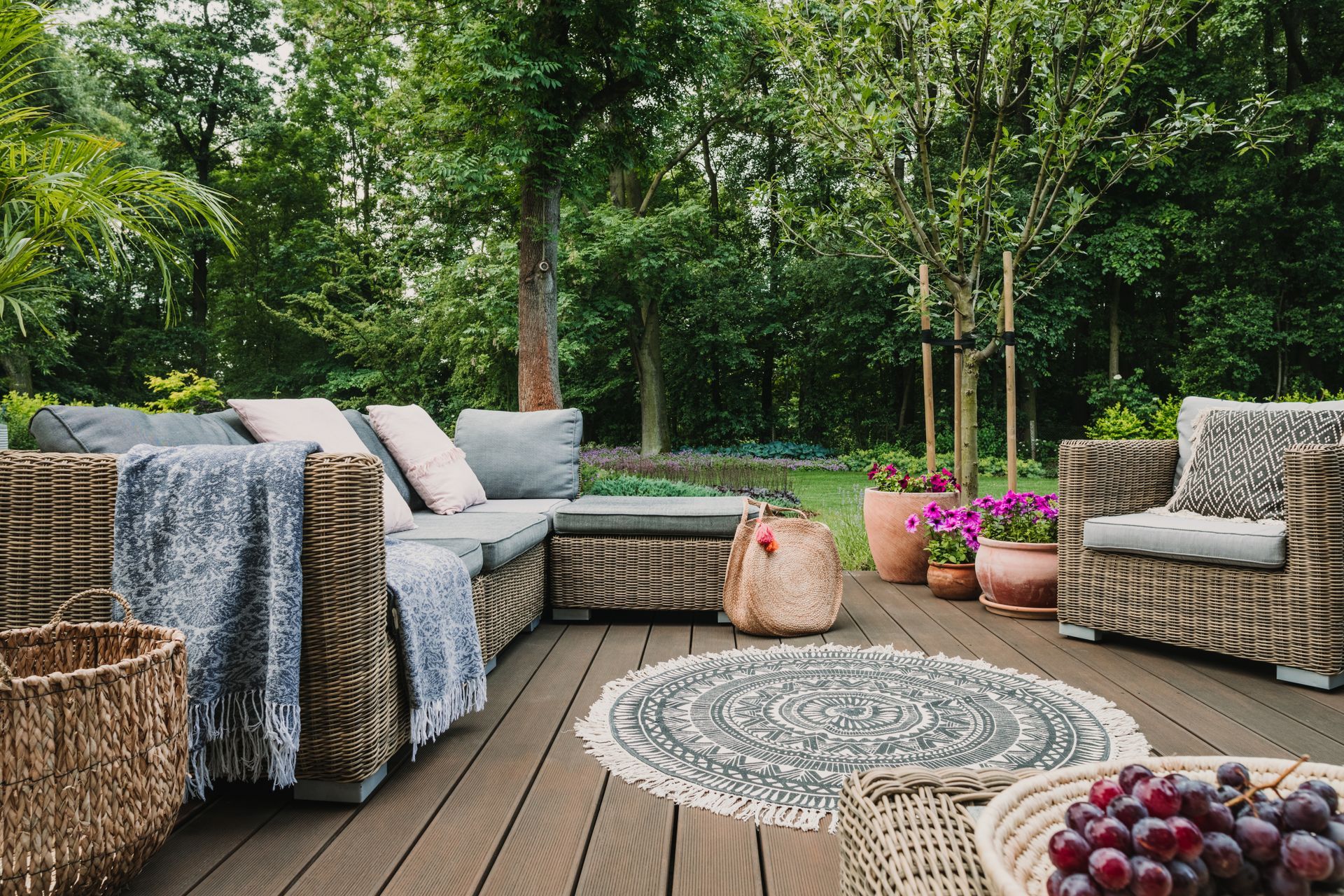 Cozy outdoor patio with wicker seating, patterned rug, potted flowers, and lush green garden backdrop