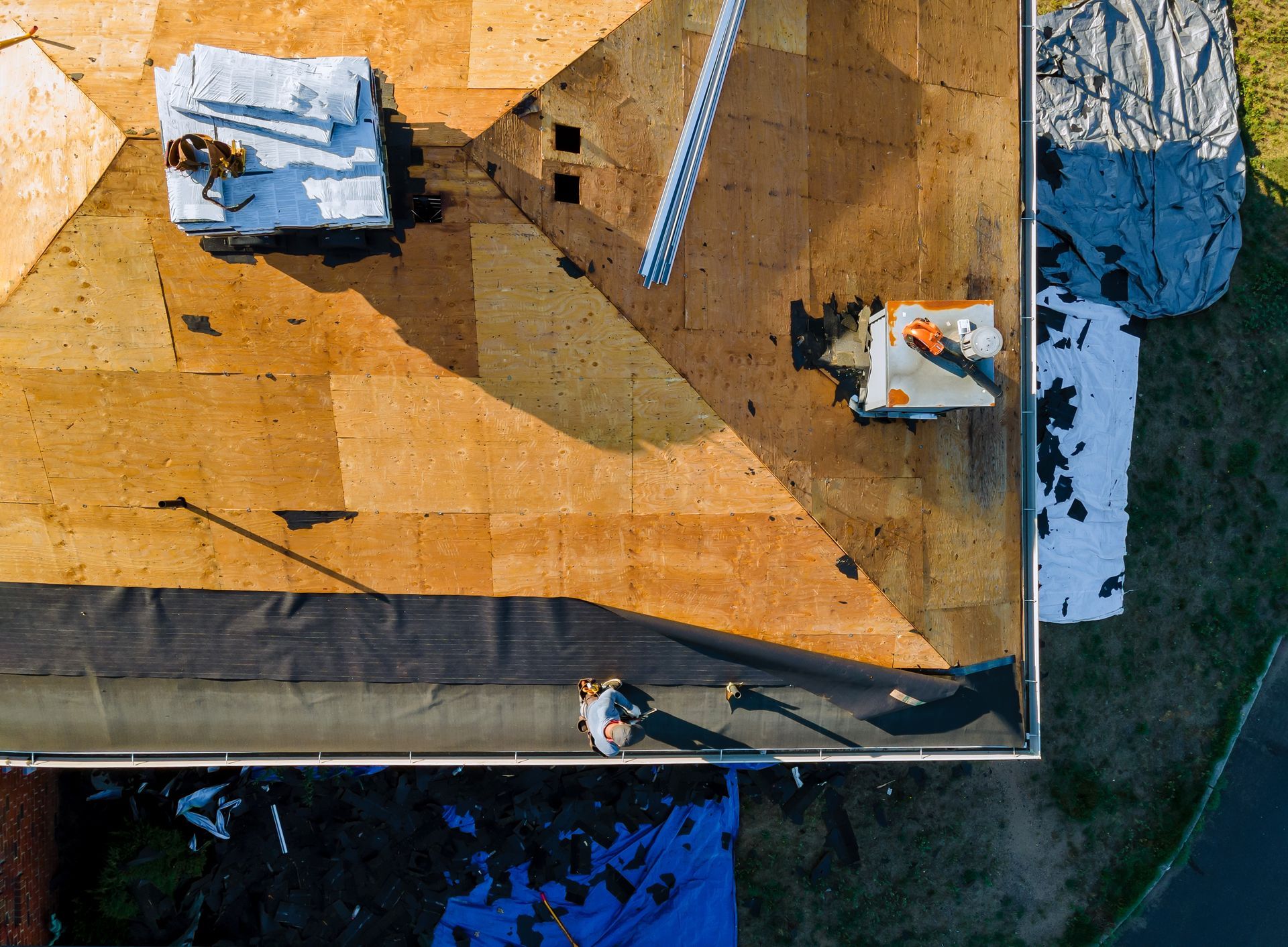 An aerial view of workers installing black underlayment on a plywood roof deck with construction materials nearby.