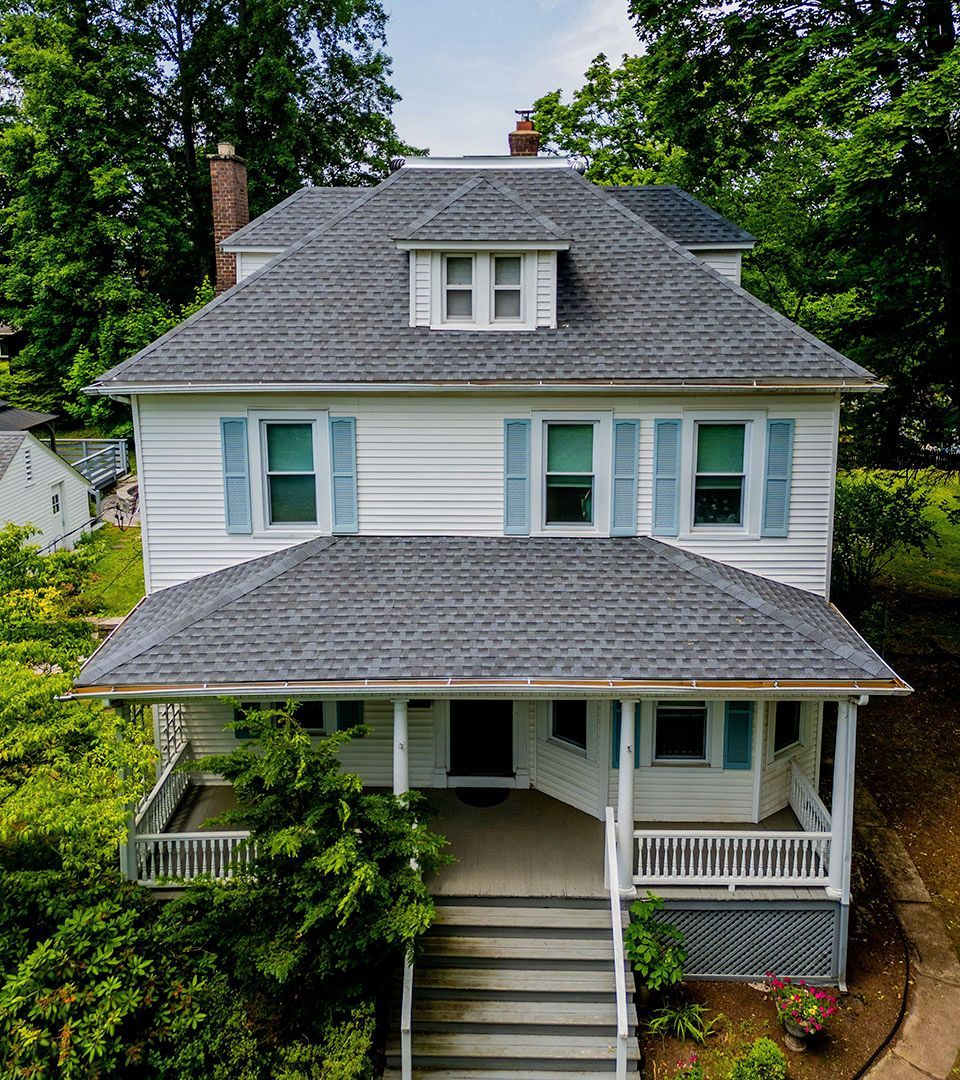 White house with blue shutters, a porch, gray roof, and surrounded by trees.