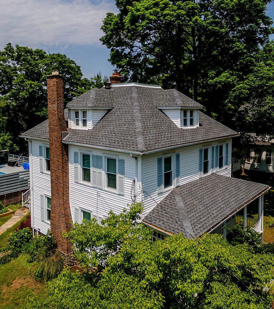 White house with a gray roof and brick chimney. Blue shutters and dormer windows. Surrounded by trees.