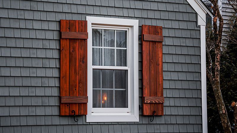 Window with white frame and dark red shutters on a gray shingled house exterior.