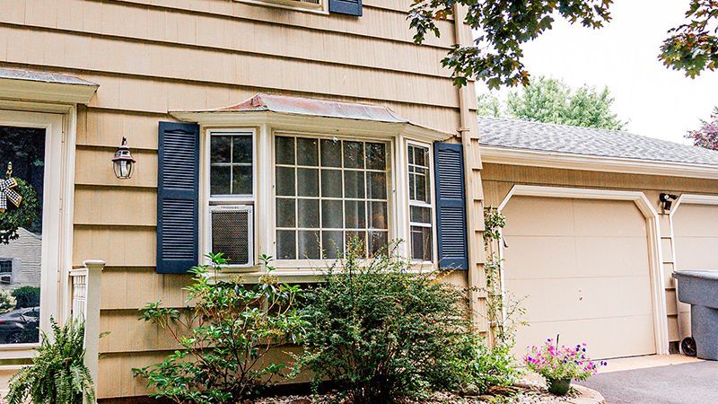 Tan house with blue shutters, bay window, and attached garage. Green bushes in front.