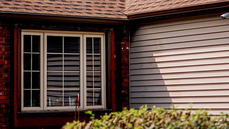 Window framed in red brick, adjacent to beige siding under a brown roof.