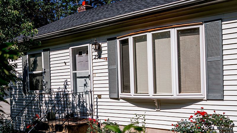 White house with dark gray shutters and bay window, front door, and dark roof.