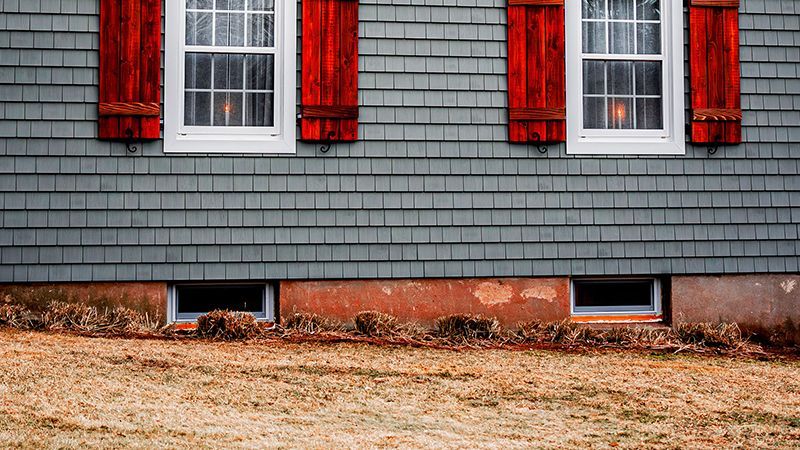 House exterior with gray siding, red shutters, and two windows. Brown ground and two basement windows visible.