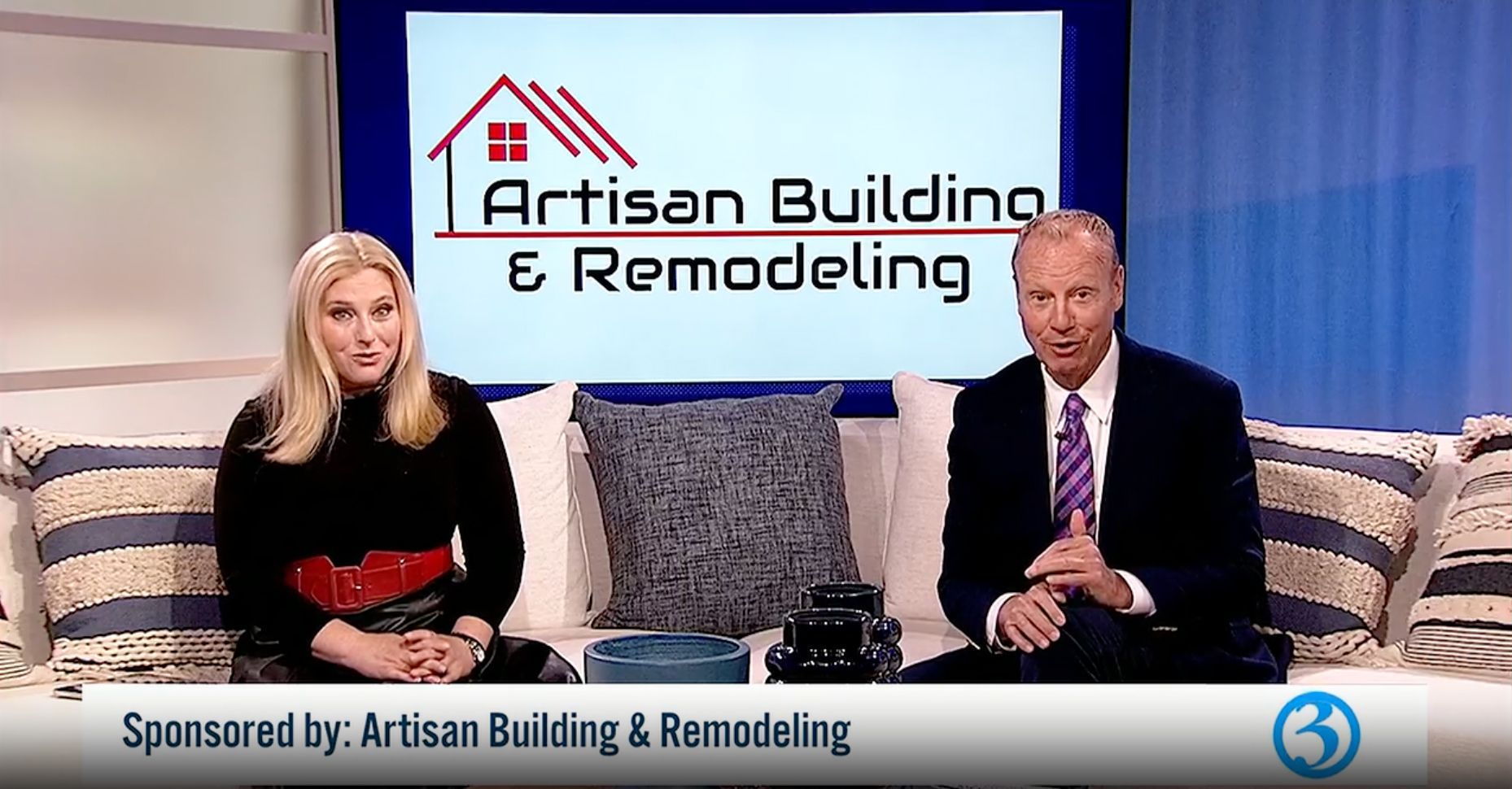 Two anchors at a desk on a studio set with an Artisan Building & Remodeling sponsor sign behind them