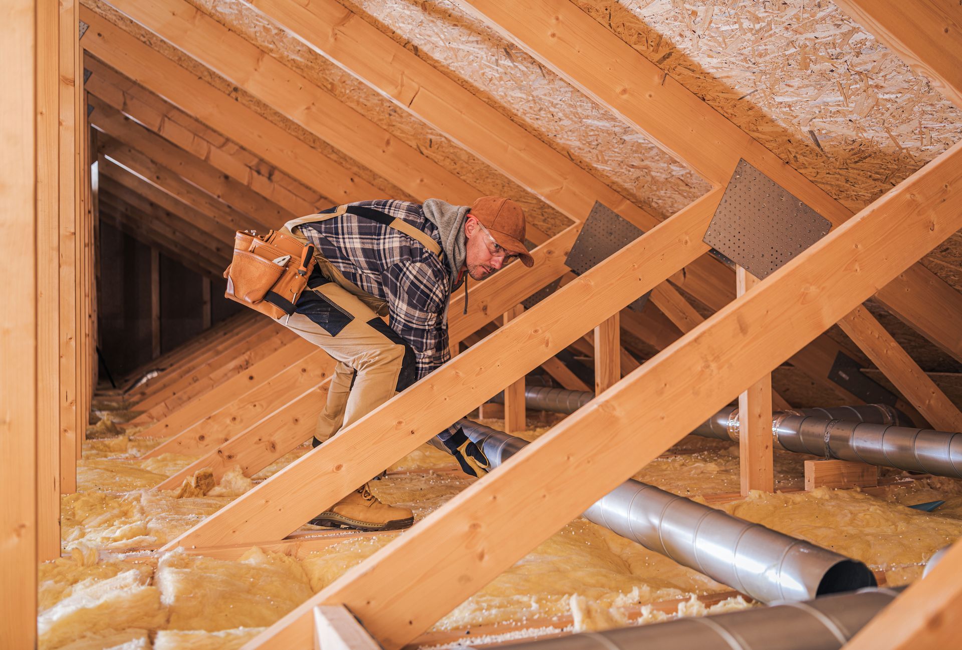 Person in an attic inspecting rafters, wearing work clothes and a tool belt.