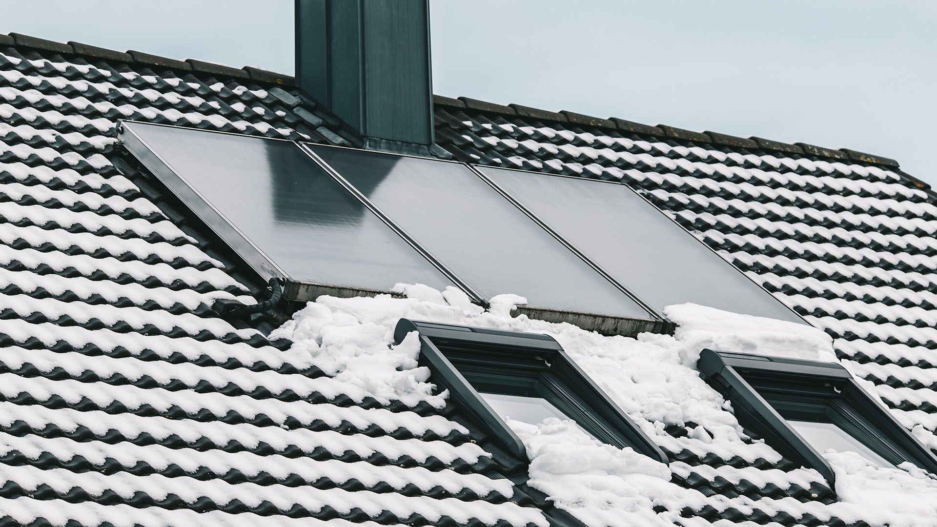 Solar panels on a snow-covered roof next to a chimney and skylights.