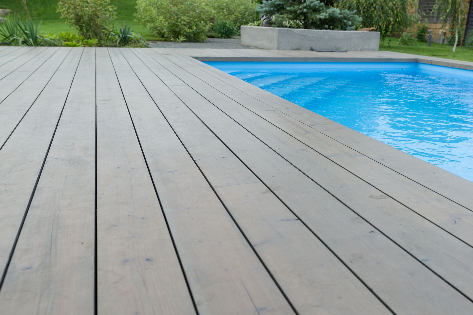Wooden deck beside a blue swimming pool with greenery in the background