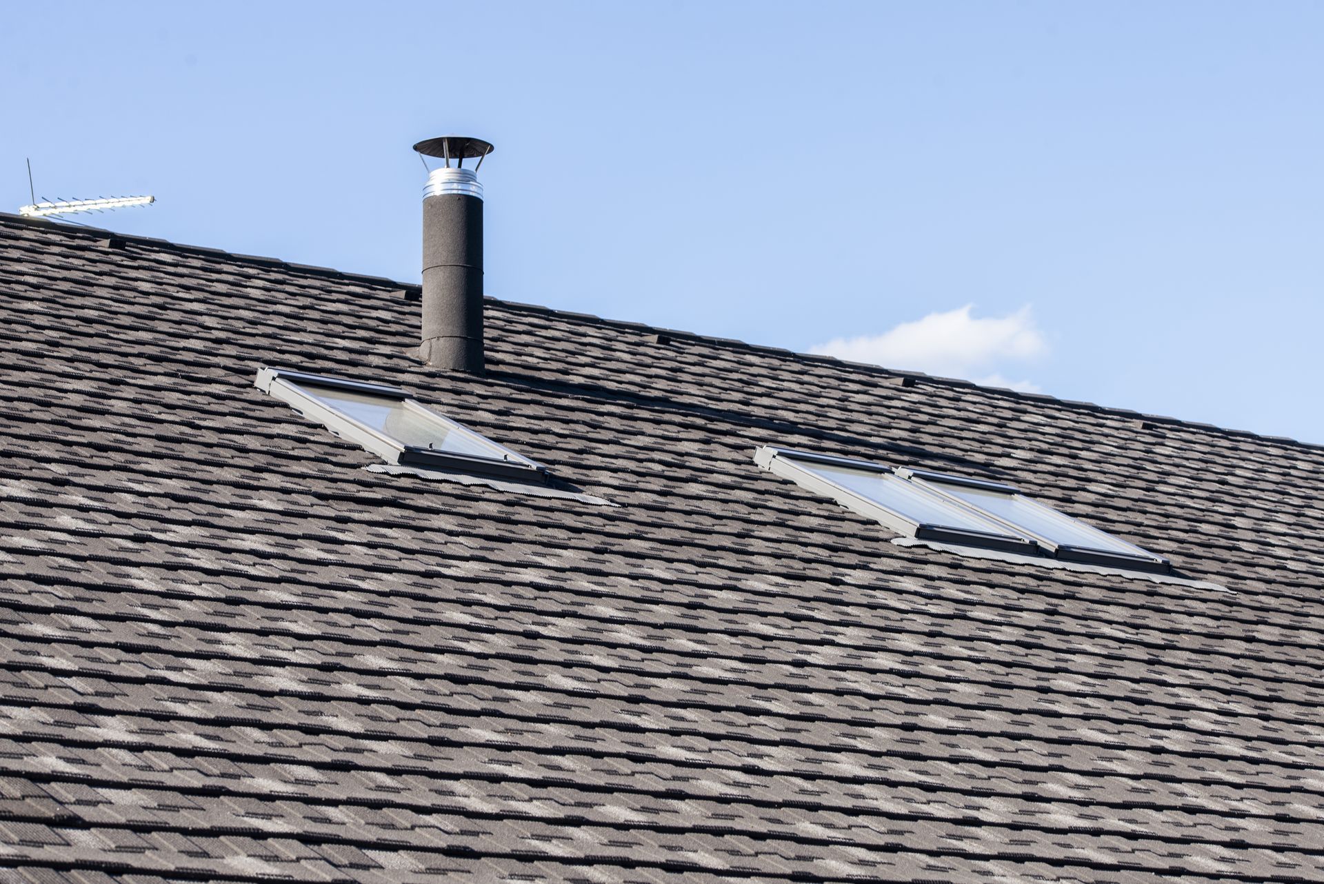 Dark shingle roof with a chimney and two rectangular vents against a blue sky.