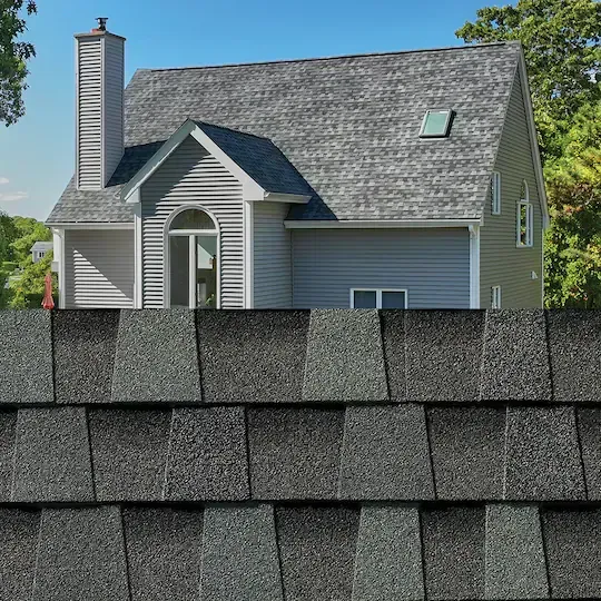 Suburban house with gray shingle roof and siding, seen above a close-up of roof shingles in the foreground