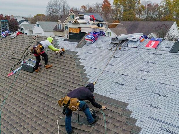 Two roofers installing shingles on a house roof. One nails, other adjusts. Overcast day.