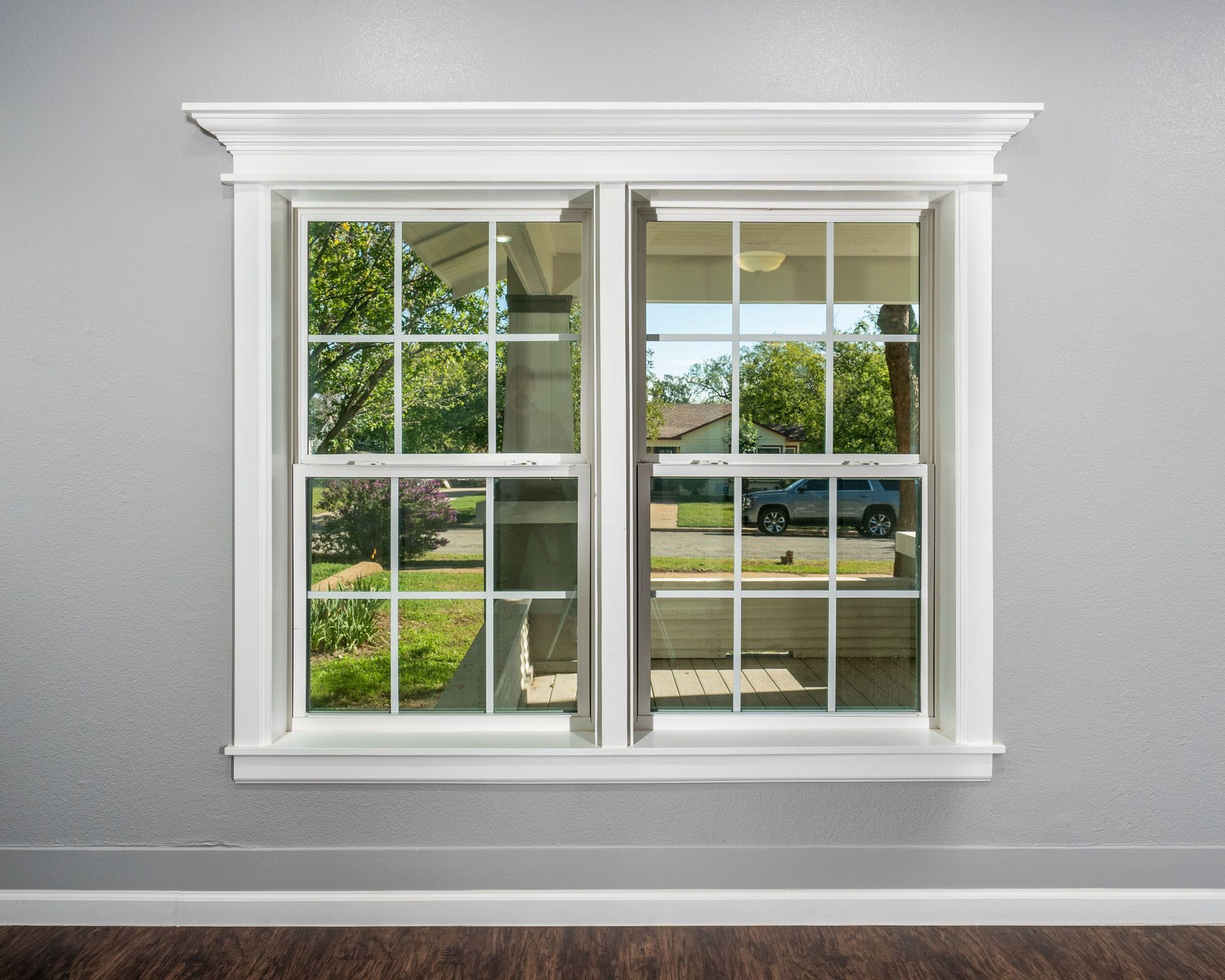 White-trimmed double window on a gray wall, reflecting a green yard and street outside.