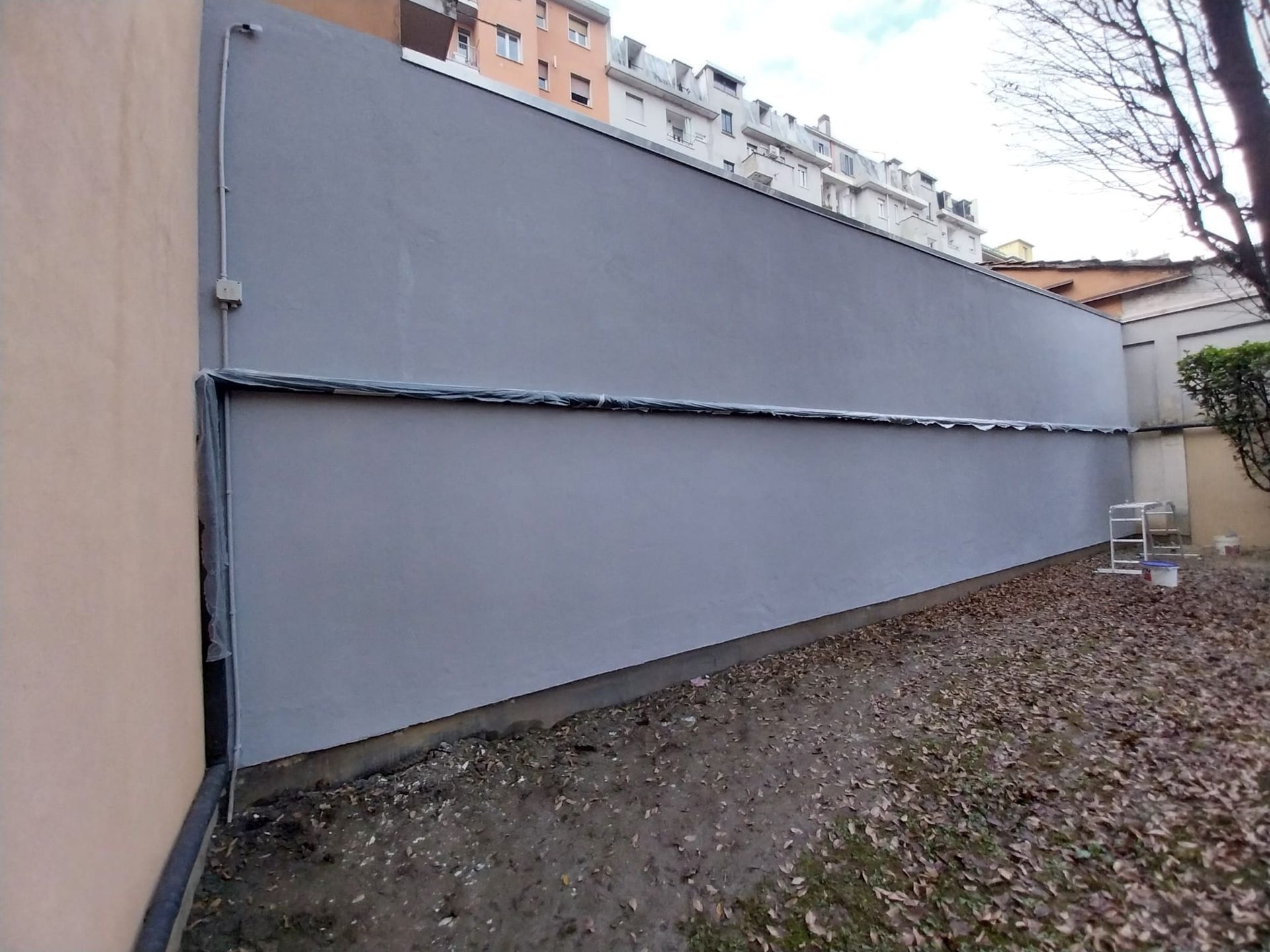 Muro grigio lungo un cortile, con giuntura visibile. Terreno coperto di foglie.