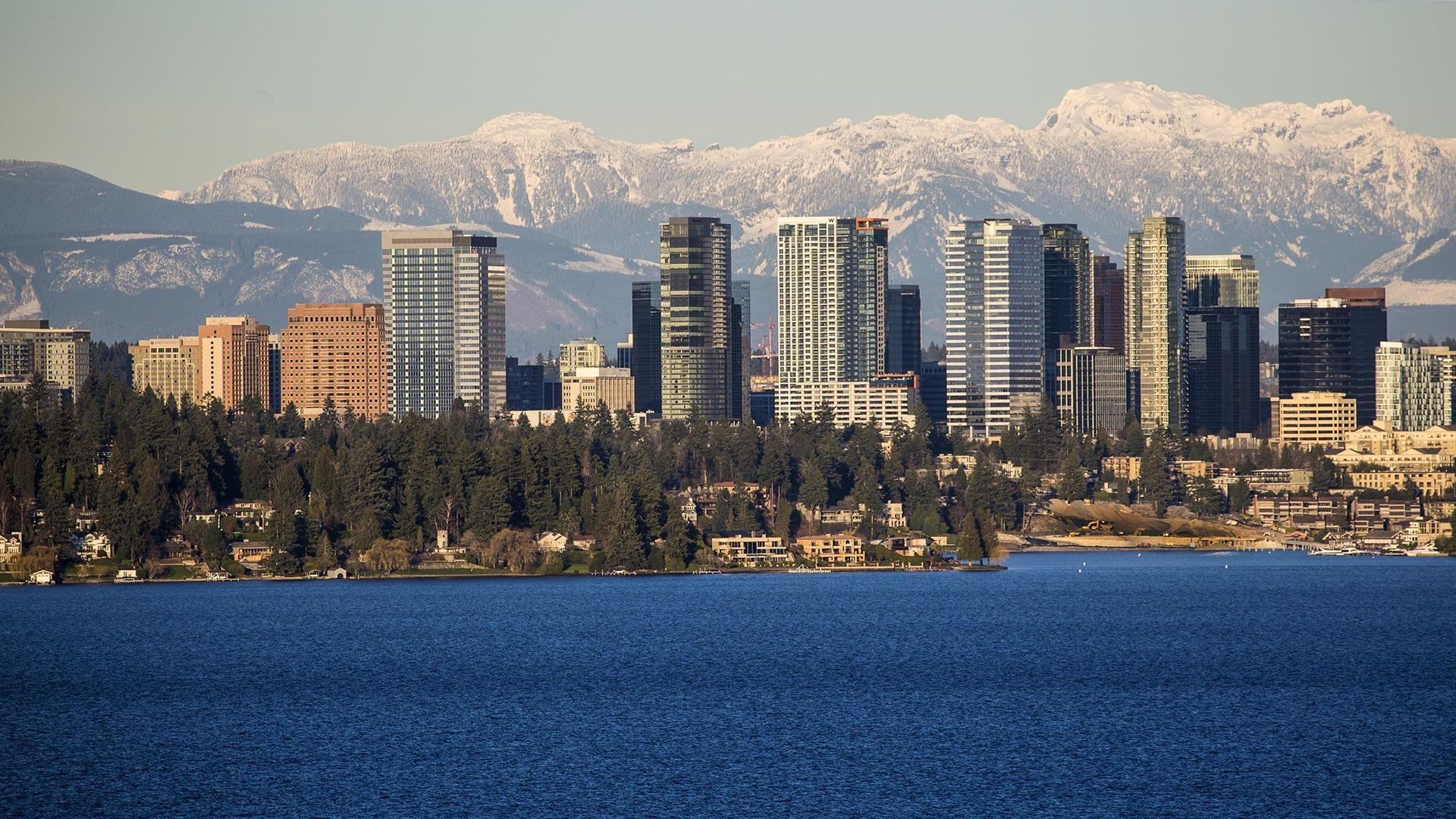 City skyline on lake, snowy mountains in background. Blue water, evergreen trees, and buildings.