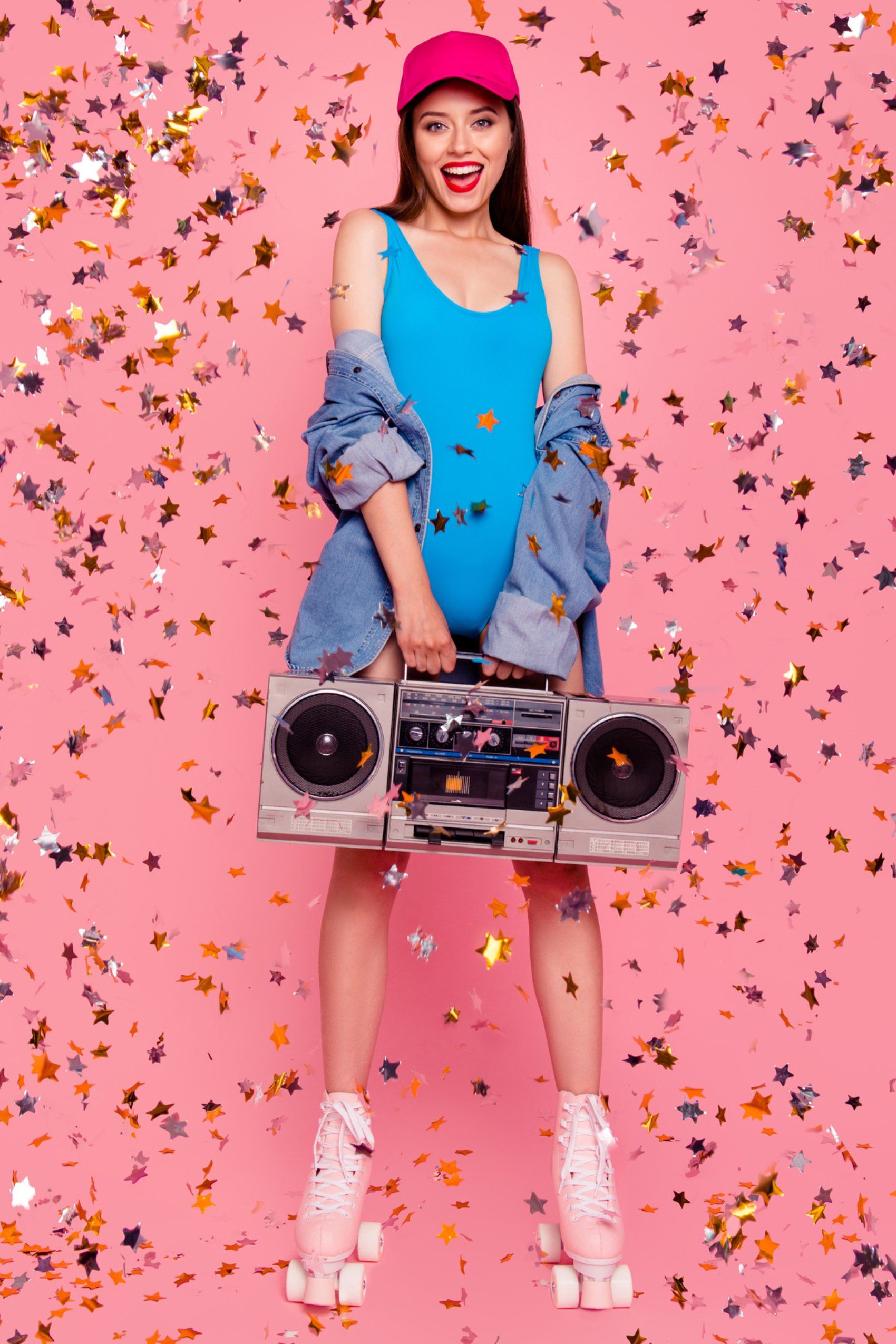 A woman in roller skates ready for a party is holding a boombox on a pink background.