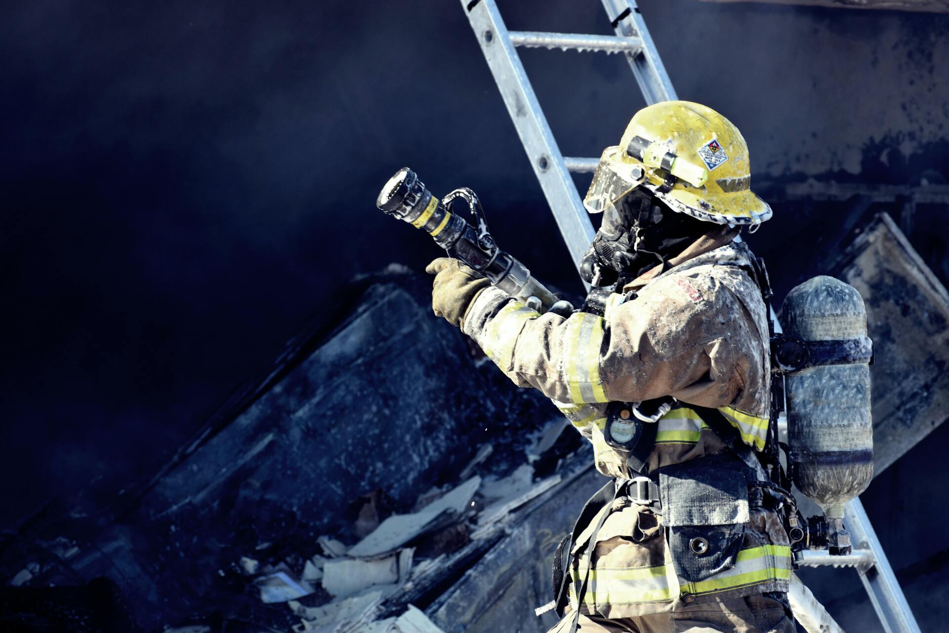 Firefighter in yellow gear spraying water at a smoky blaze beside a ladder and debris