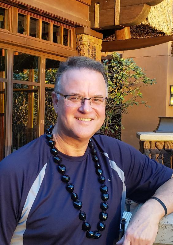 Smiling person in a navy shirt and black beaded necklace, standing outdoors by a wooden building.