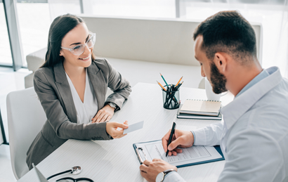 Two people in a bright office discussing paperwork at a desk with a clipboard and pens.