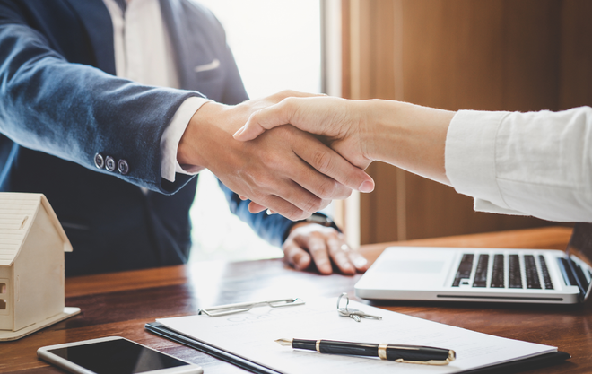 Business handshake over desk with laptop, clipboard, and pen in a bright office