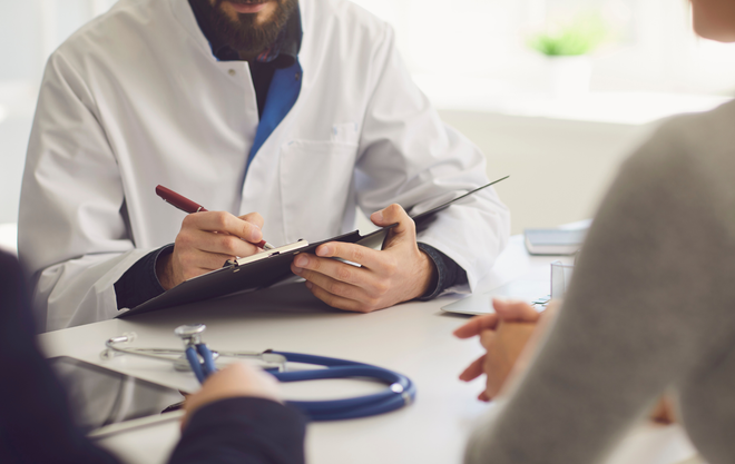 Doctor writing on a clipboard during a consultation, with a stethoscope on the table.