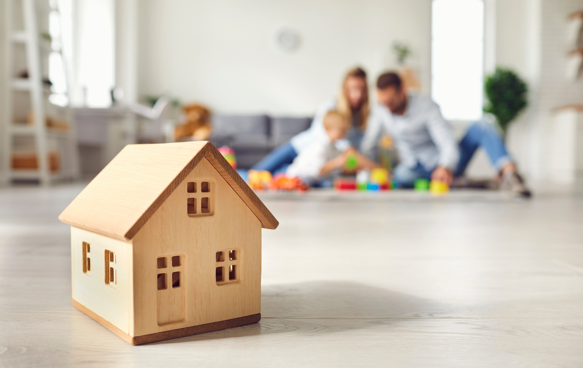 Small wooden house model on a table, with a blurred family playing in the background.