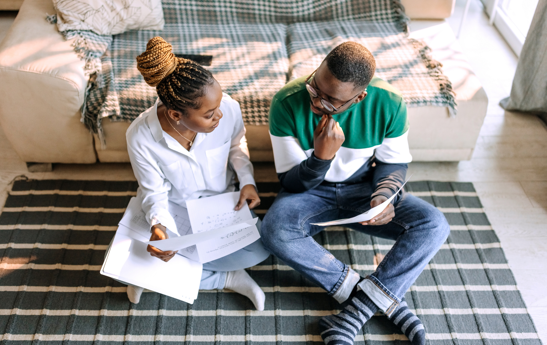 Two people sit on a living room rug, reviewing papers and talking near a sofa.
